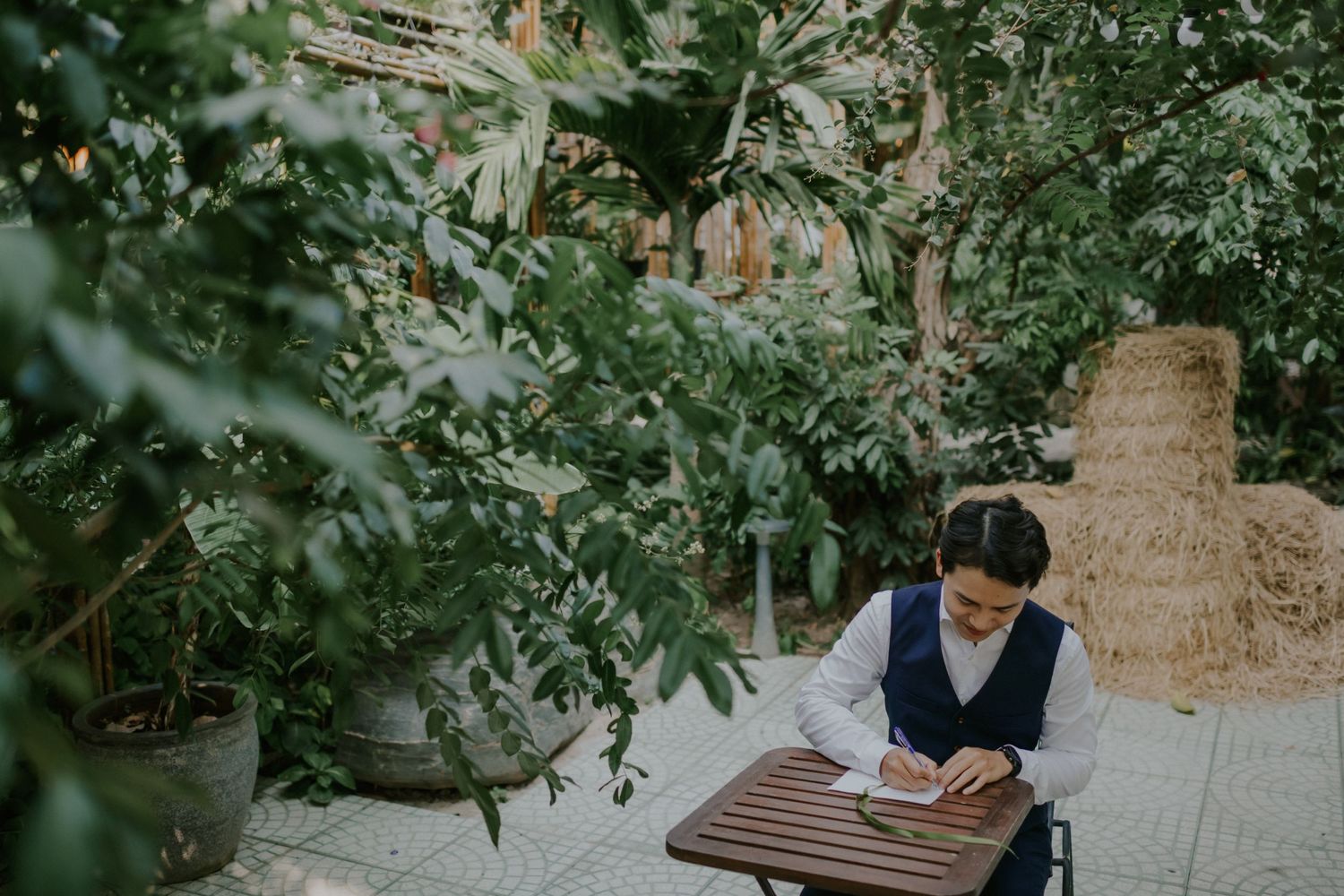 Person in formal attire sits at outdoor table writing in notebook surrounded by greenery.