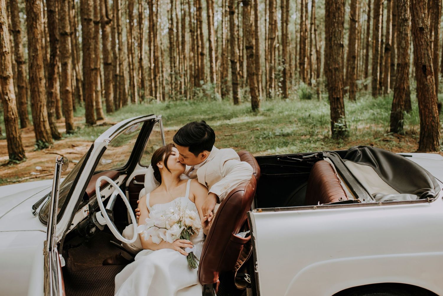 A romantic moment shared in a vintage white convertible surrounded by tall pine trees.