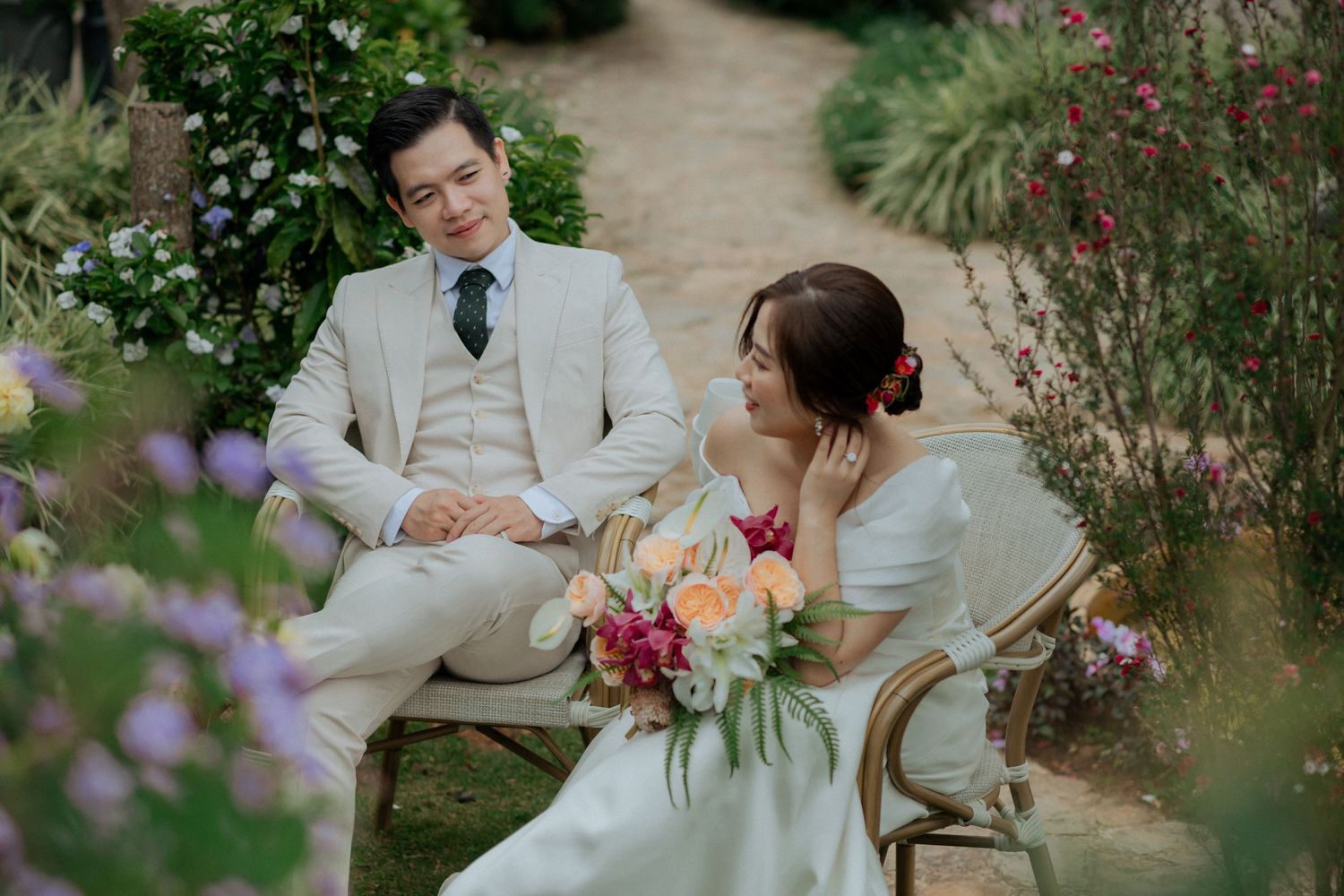 Wedding couple sits on vintage chairs in a lush garden path with pink and purple flowers blooming around them.
