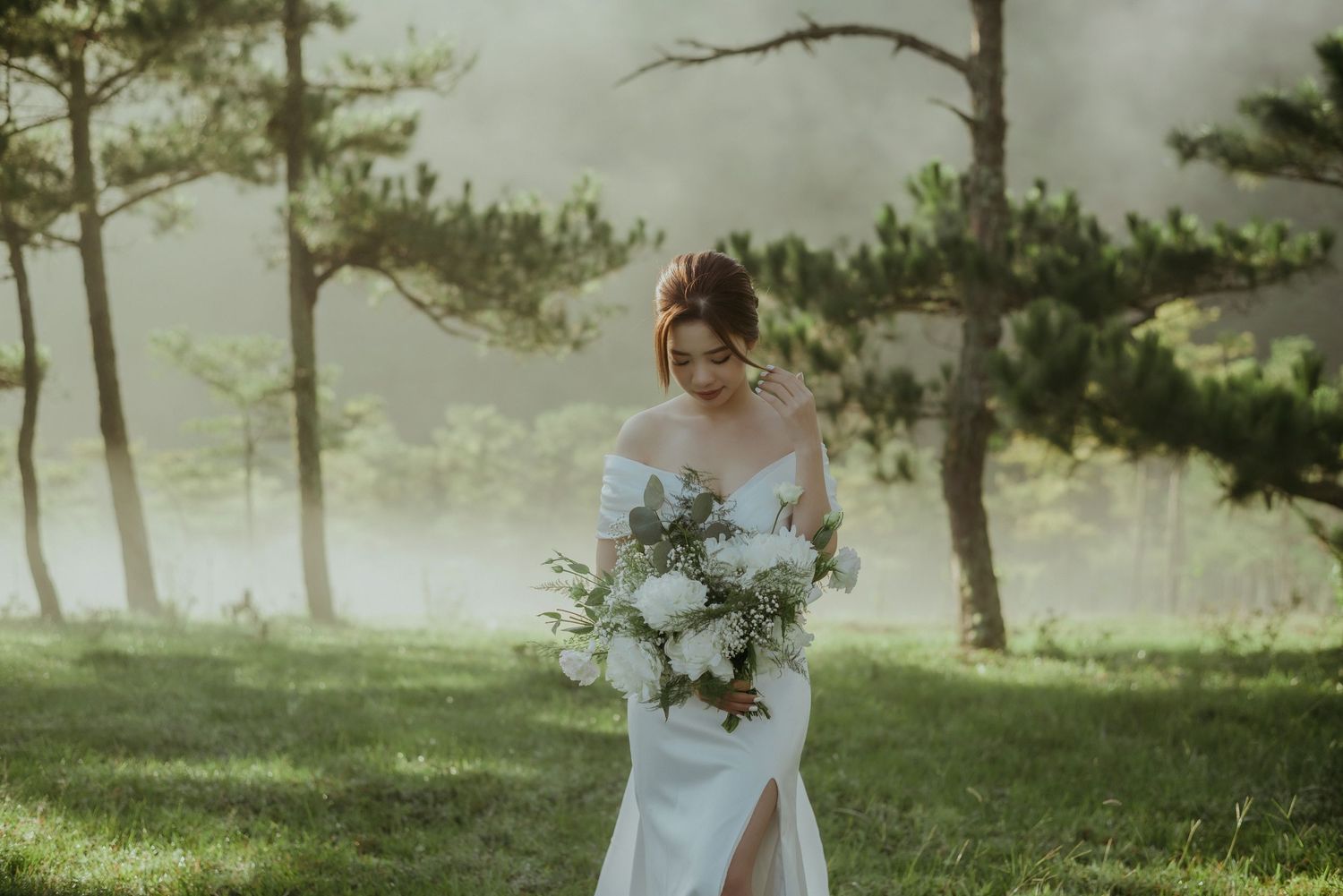 Person in white wedding dress holding elegant white floral bouquet poses among misty pine trees on grassy slope.