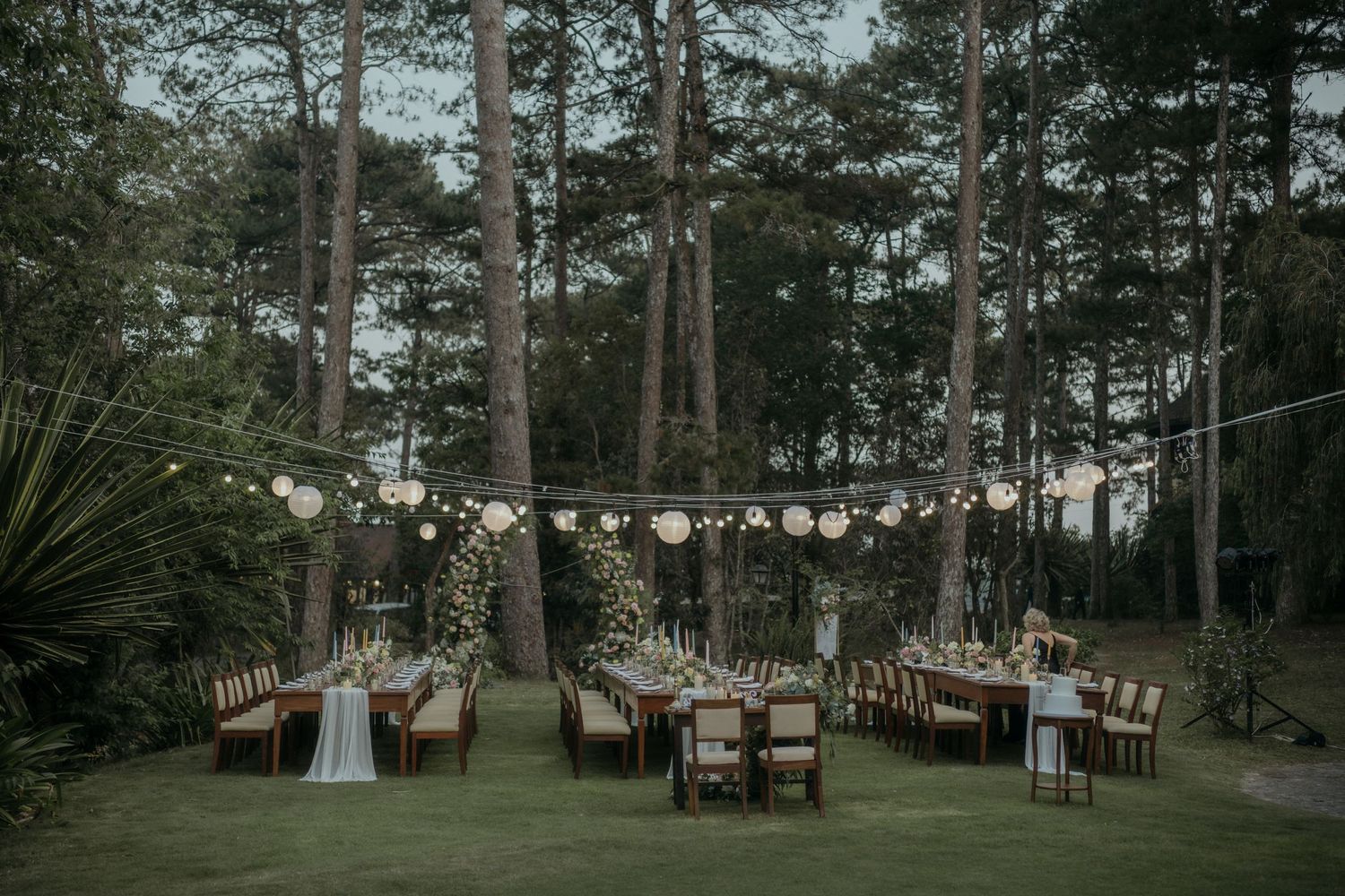 Rustic outdoor wedding reception setup in a forest with string lights and wooden tables at dusk.