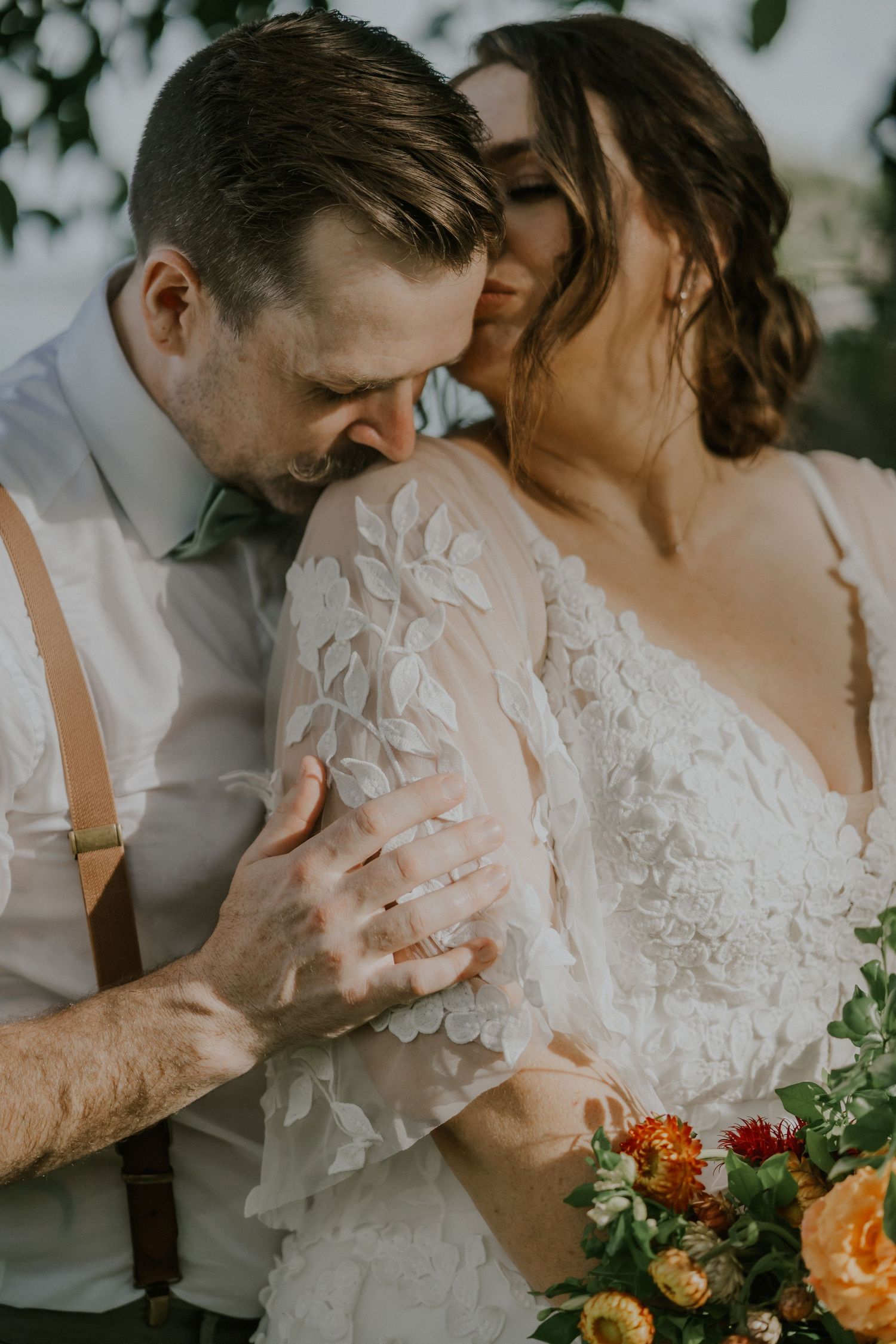 Tender wedding moment captured as couple embraces under floral decorations with orange bouquet.