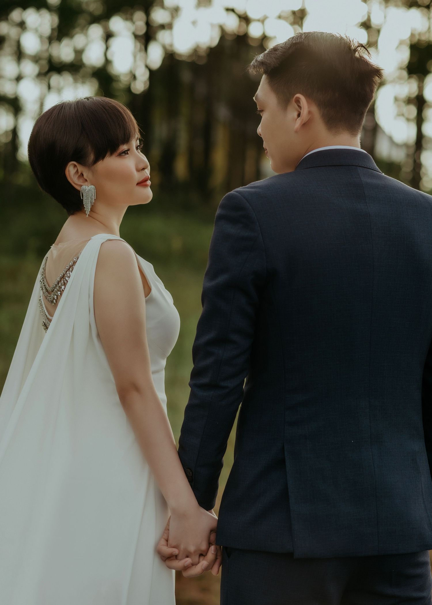 Couple in wedding attire holding hands while standing together in a sunlit forest setting.