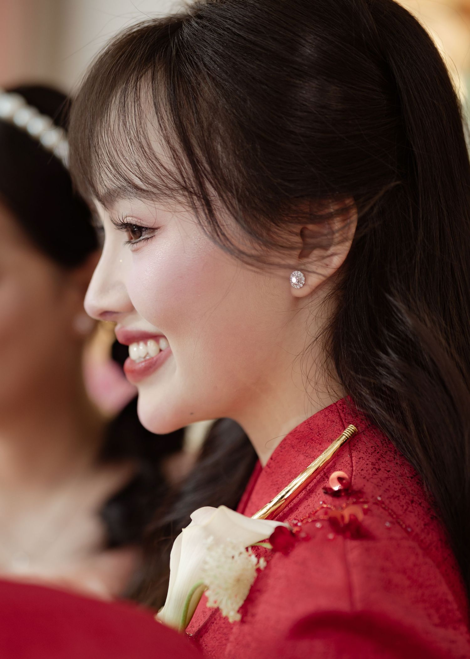 Side profile view of someone with dark hair and a red dress smiling while wearing delicate pearl earrings.