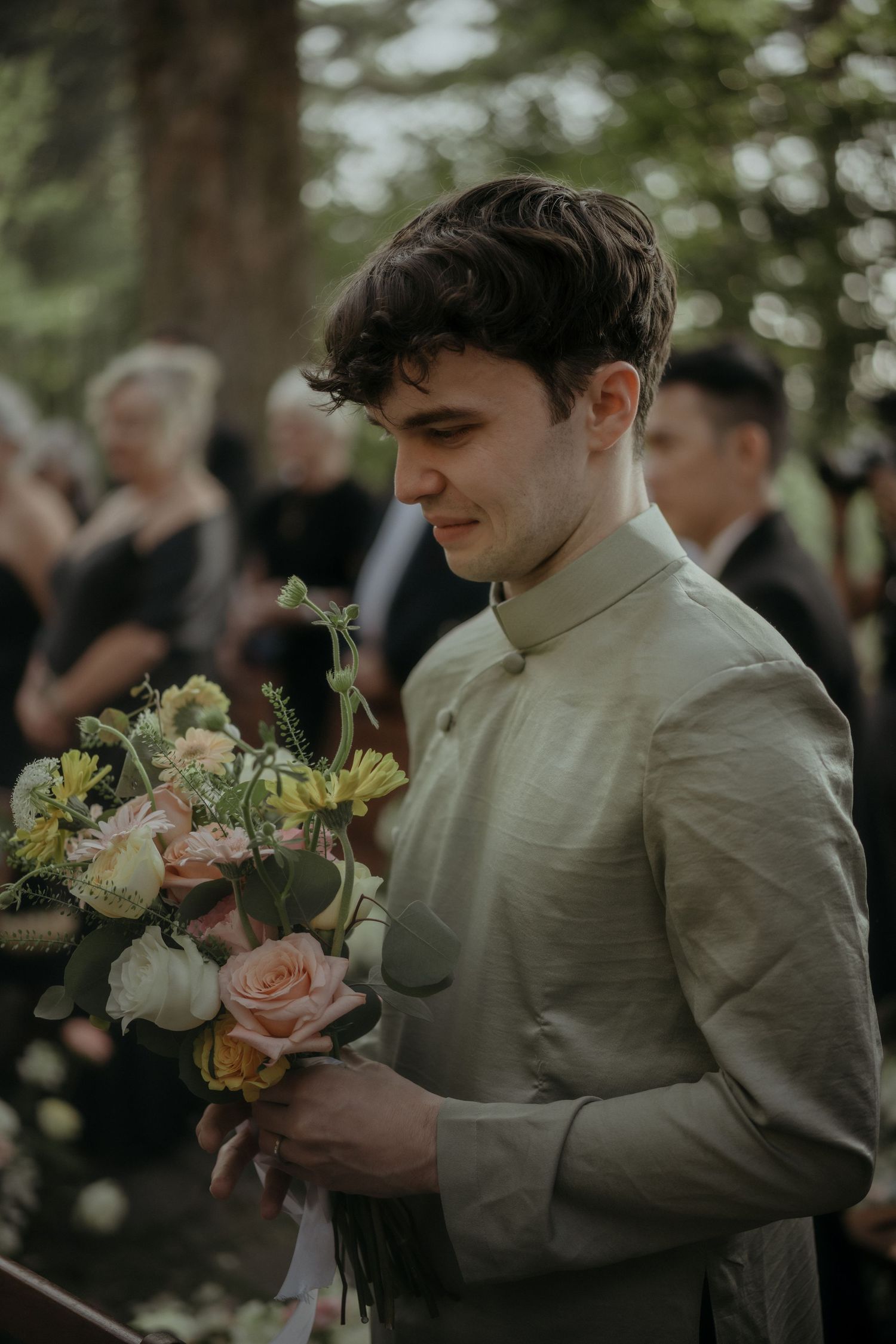 A person in a light colored shirt holds a delicate bouquet of roses and flowers at an outdoor event.