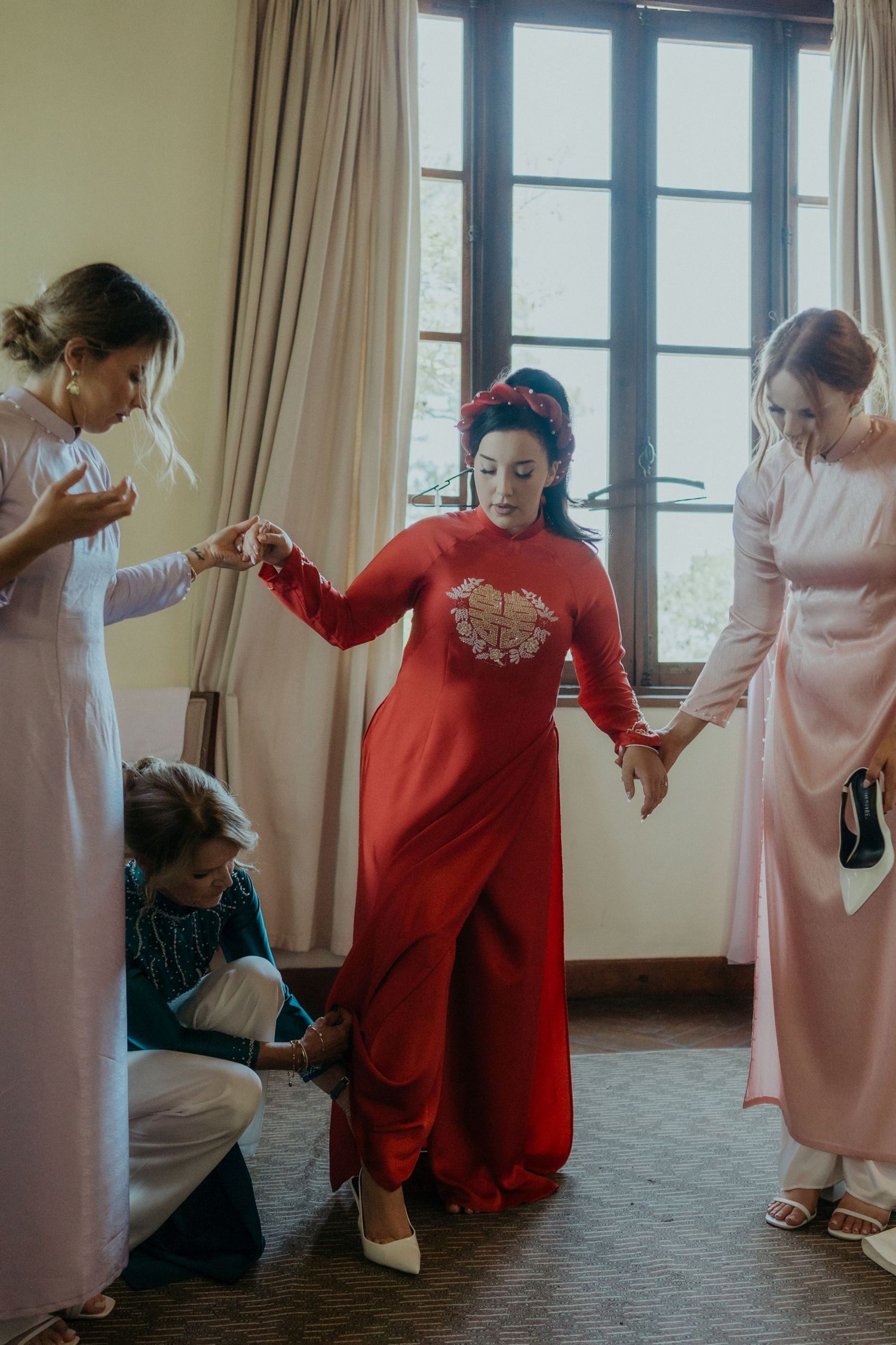 A group wearing traditional formal attire assist someone in a red qipao dress during what appears to be a wedding preparation.