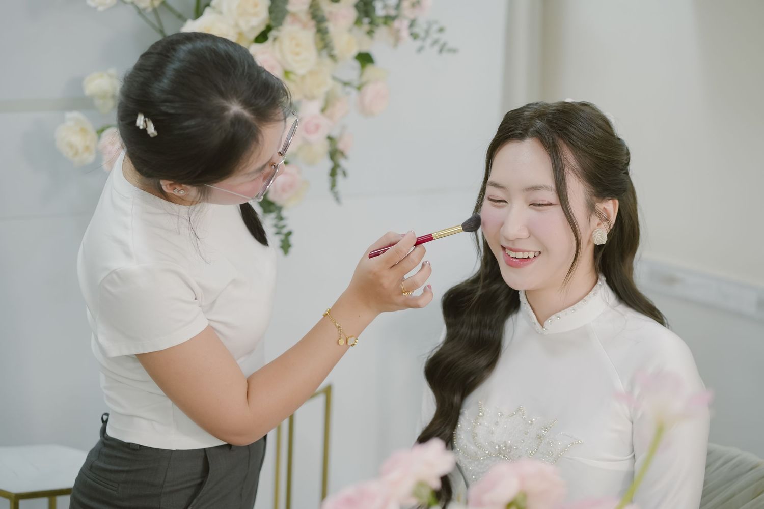 A joyful moment during bridal makeup preparation in a light-filled room.
