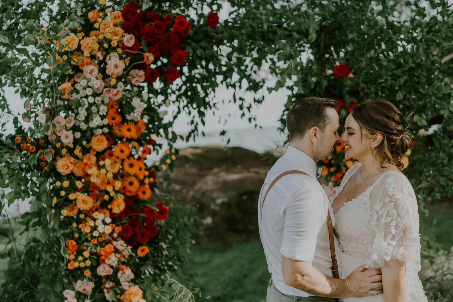 Wedding couple stands intimately under a vibrant floral arch with orange and red blooms in an outdoor garden.