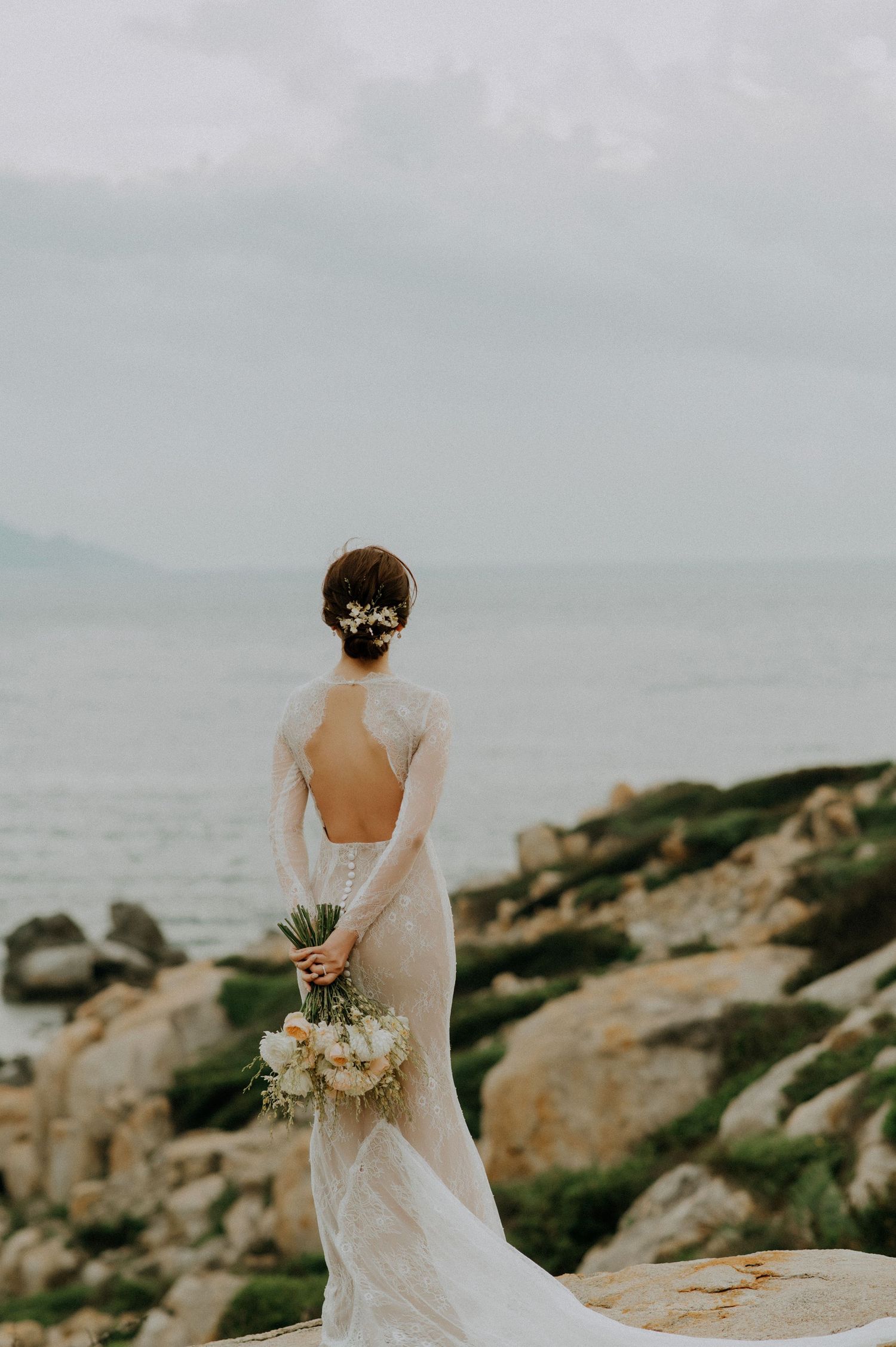 A bride in a backless lace wedding gown holds a white bouquet while standing on coastal rocks at sunset.