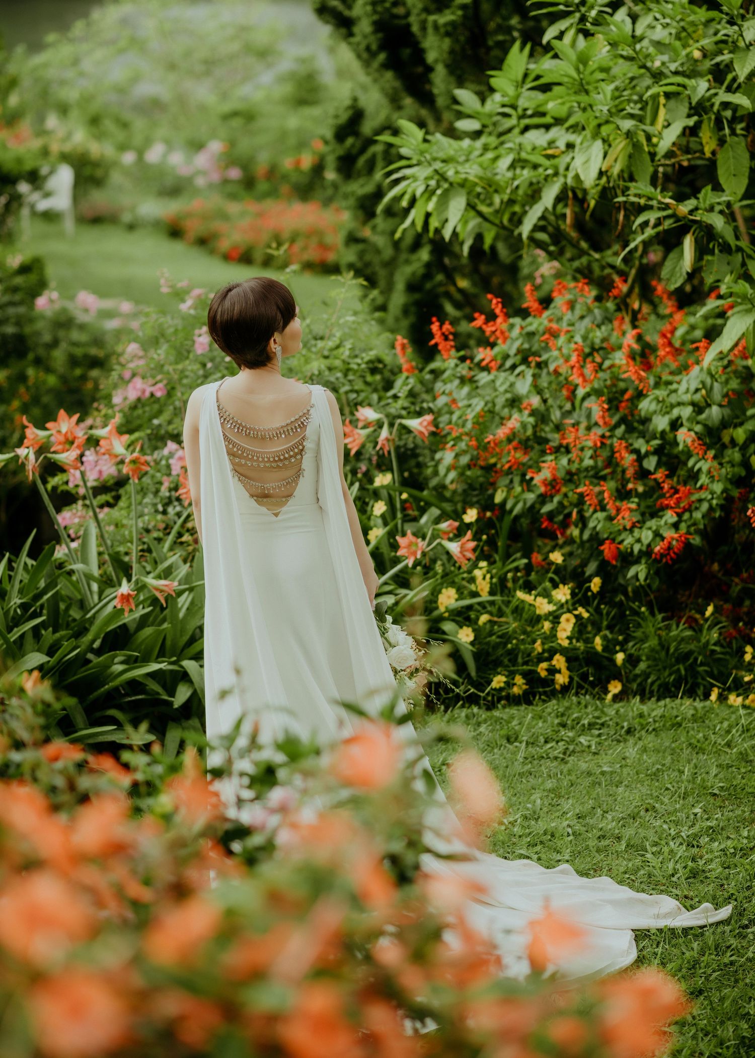 Figure in flowing white dress with open back detail stands in lush garden with blooming coral flowers and greenery.