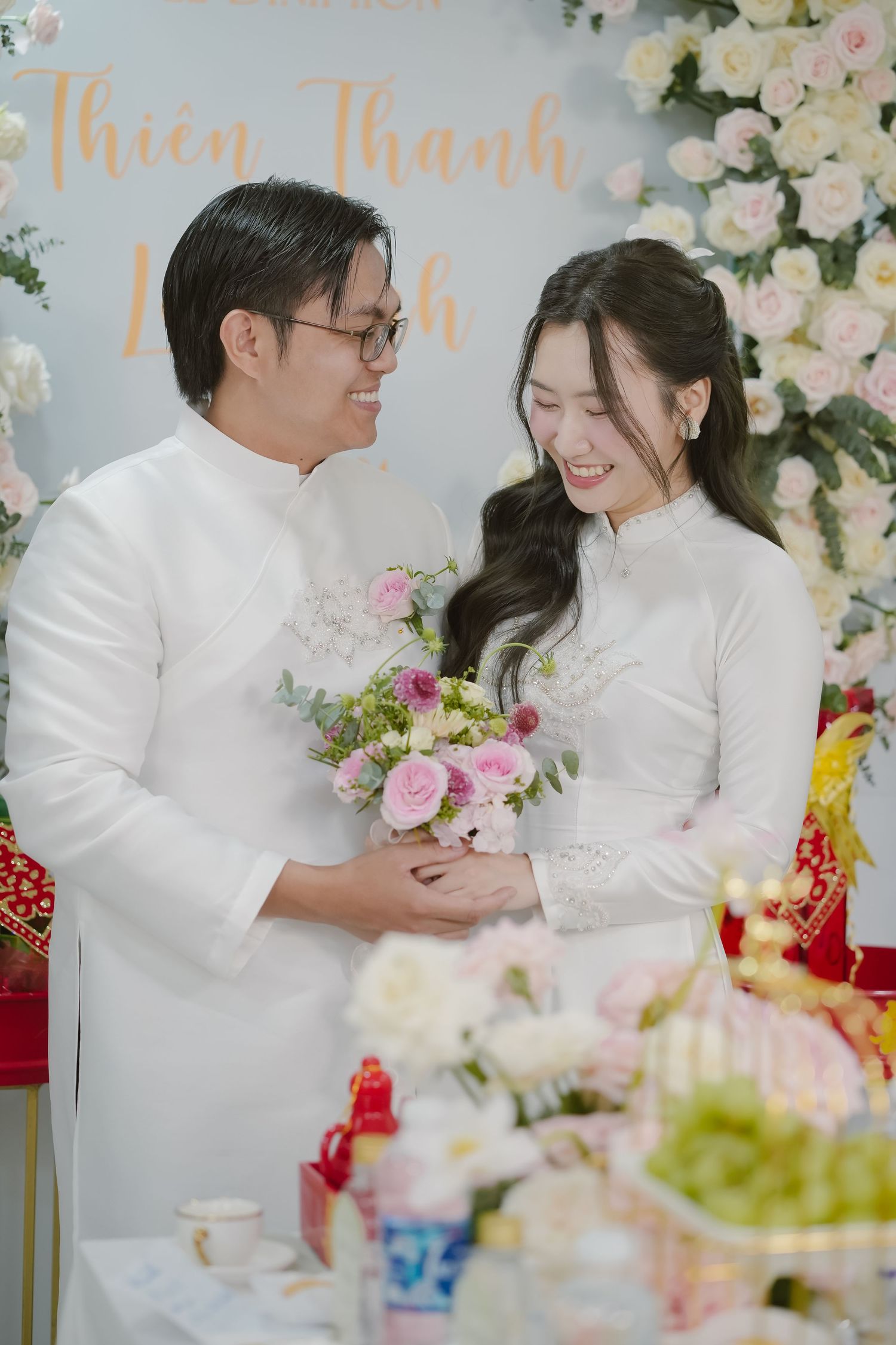 Couple in traditional white ao dai sharing moment at wedding reception with floral backdrop.