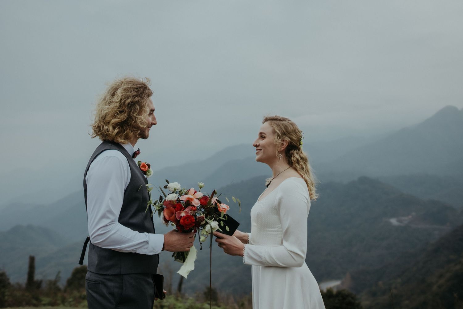 A couple shares an intimate wedding moment on a mountaintop with a bouquet of red flowers.