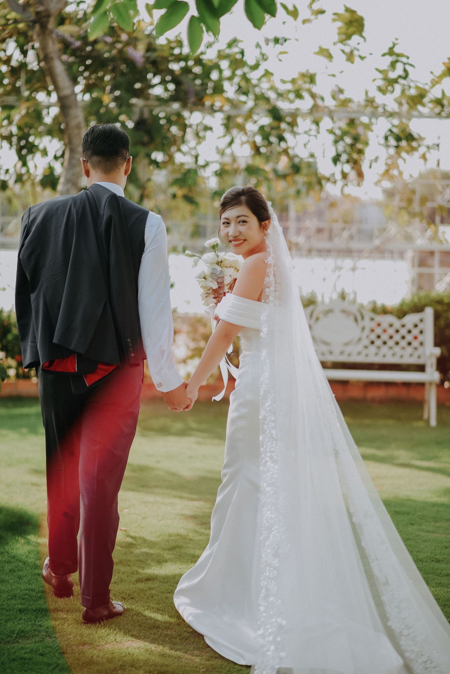Bride in flowing white dress looks back while walking with groom on garden lawn.