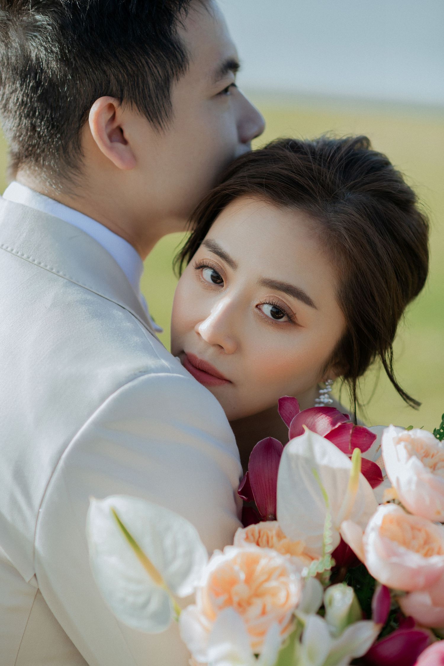 Romantic portrait with soft peach and pink flowers features a couple in white formal attire embracing outdoors.