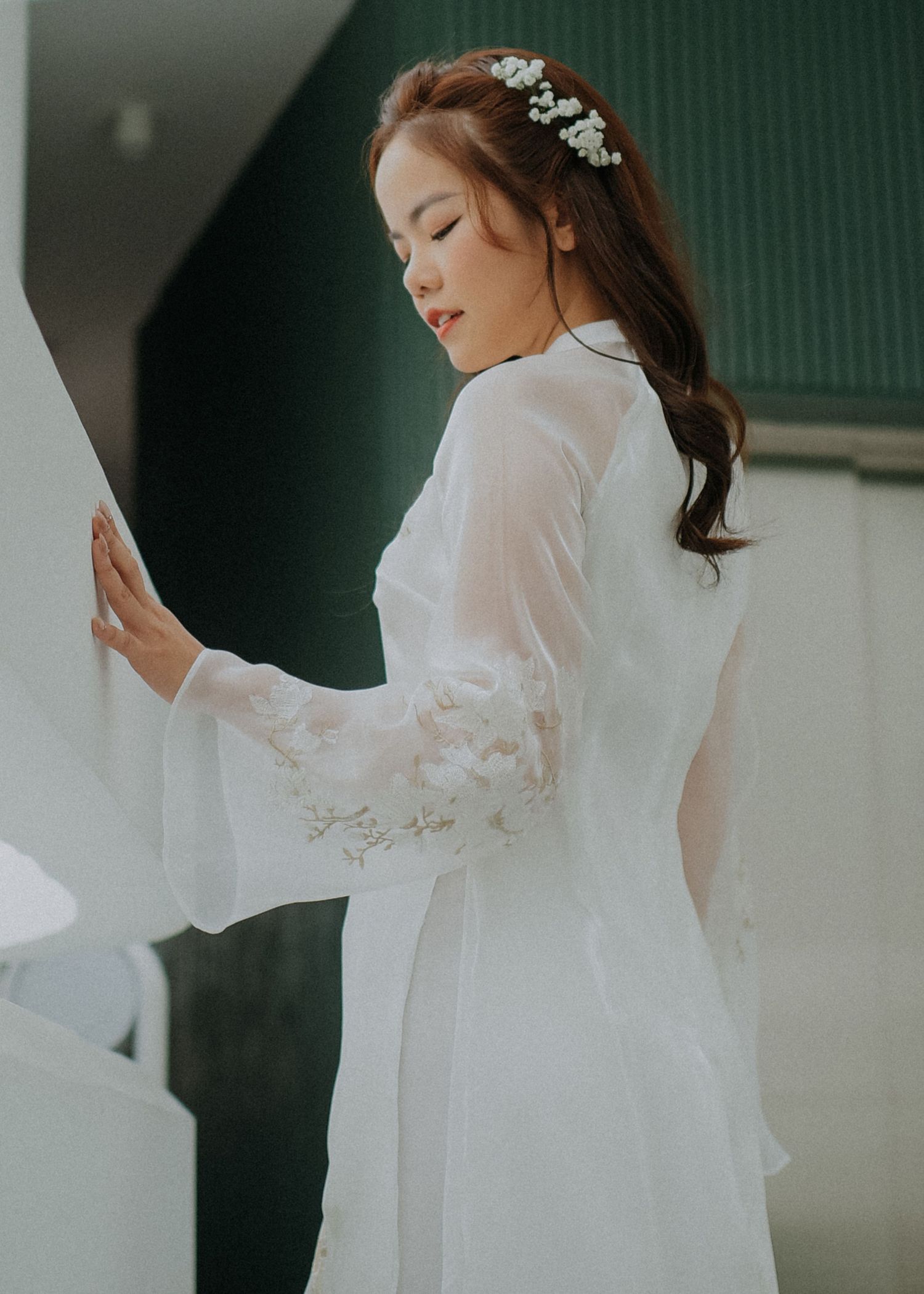 Side profile of bride in white ao dai with delicate hair accessories against green wall.