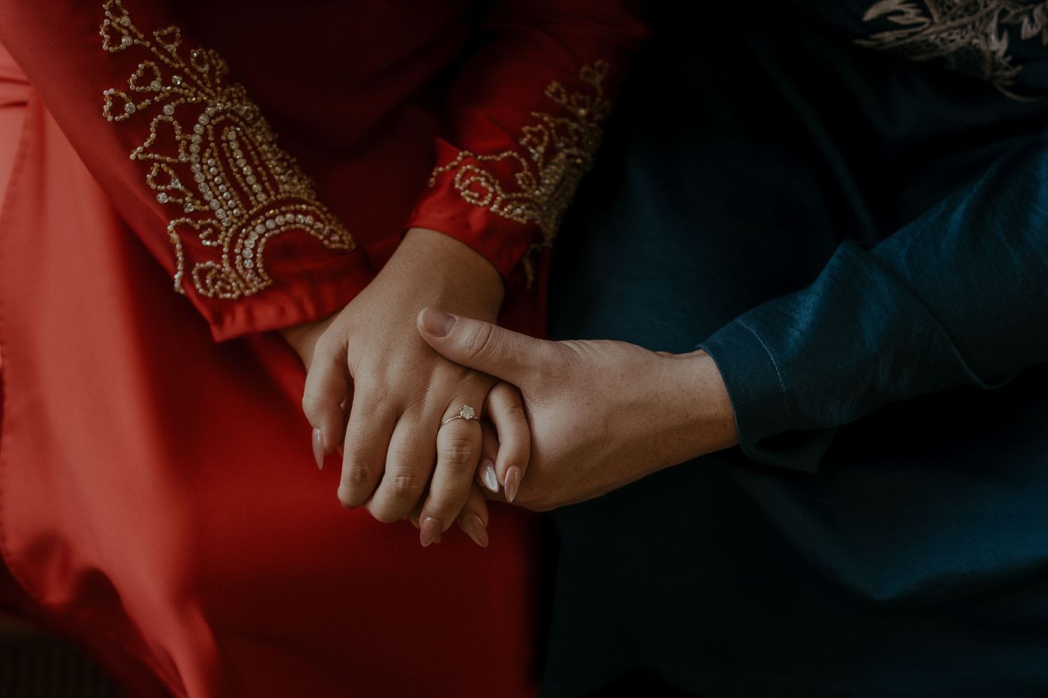 Two hands clasped together against a backdrop of red and navy blue traditional garments with gold embroidery.