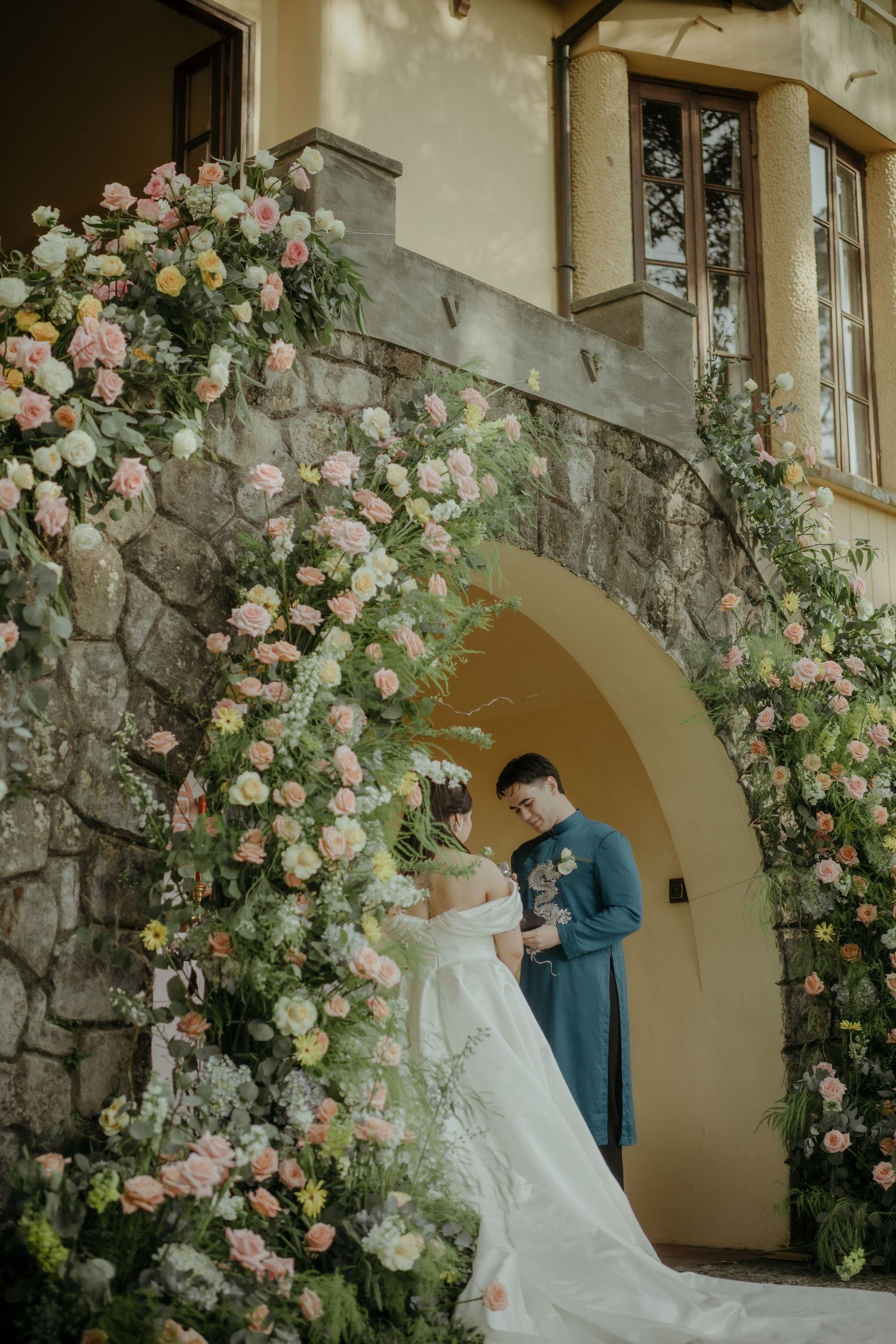 Romantic stone archway decorated with pink and cream roses framing a couple in wedding attire at elegant venue entrance.