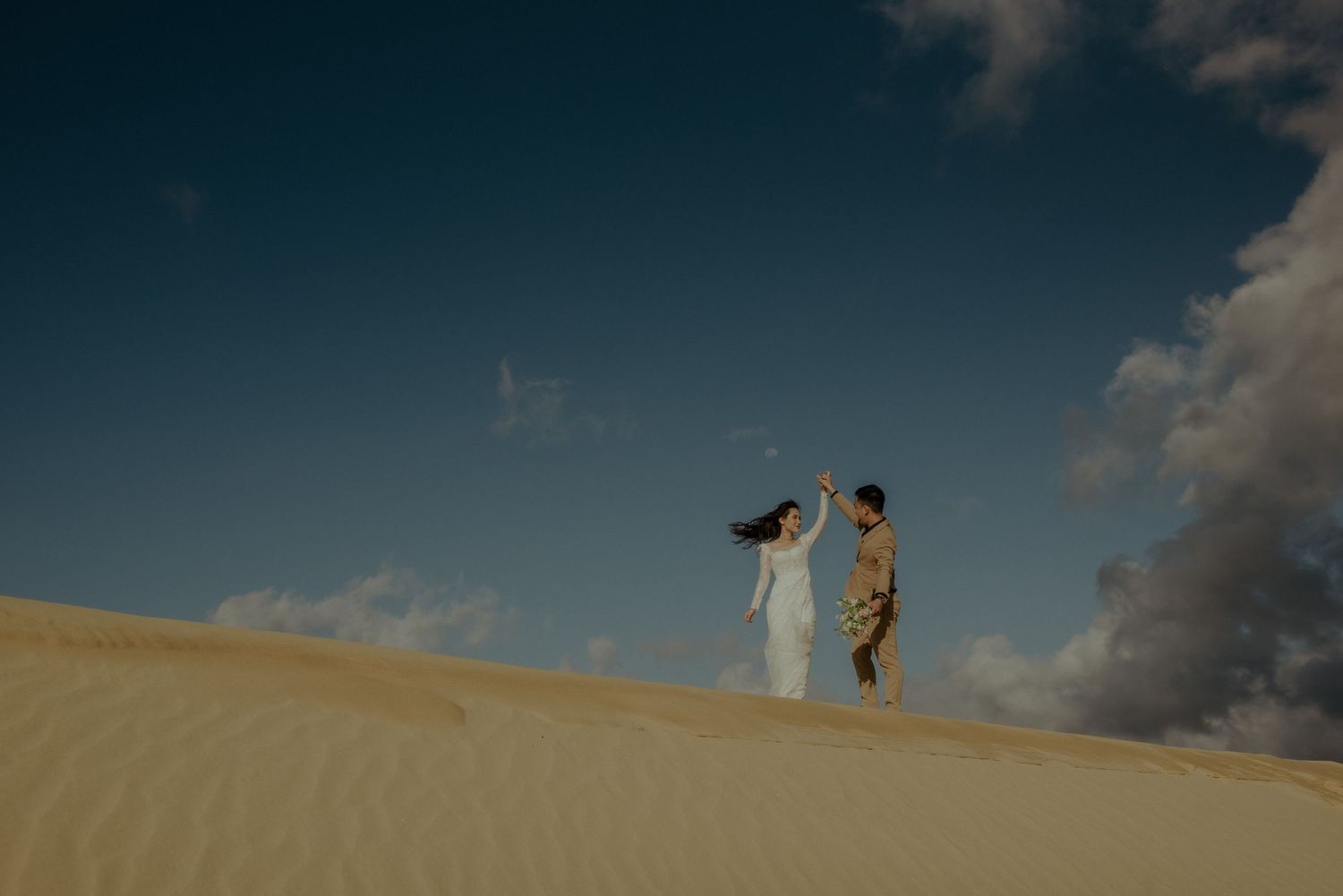 Two figures stand against a dramatic sky atop a sandy dune with flowing fabric catching the desert breeze.