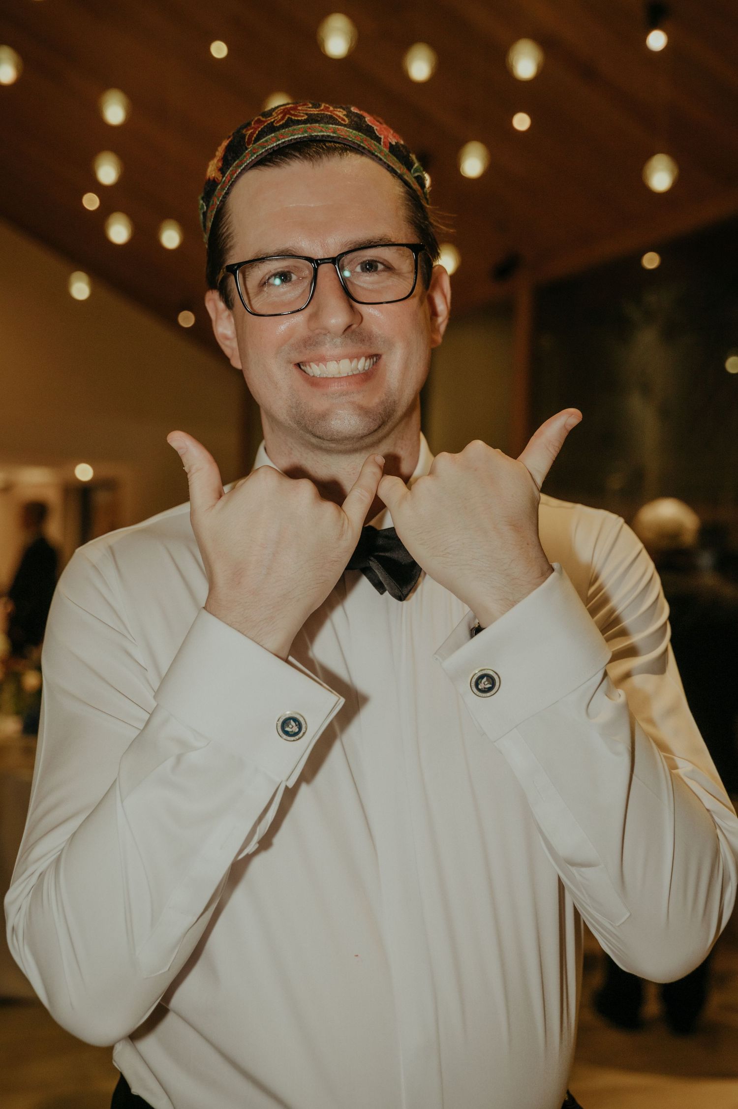 Person in a white dress shirt and bowtie making playful hand gestures under string lights.