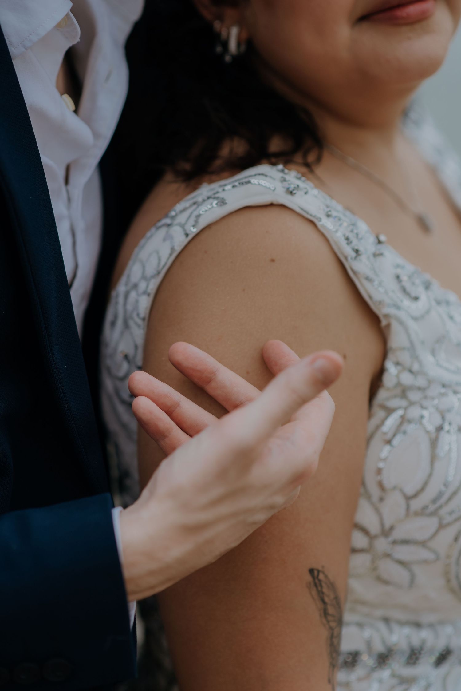 Close-up detail of white lace wedding dress and gentle touch on shoulder.