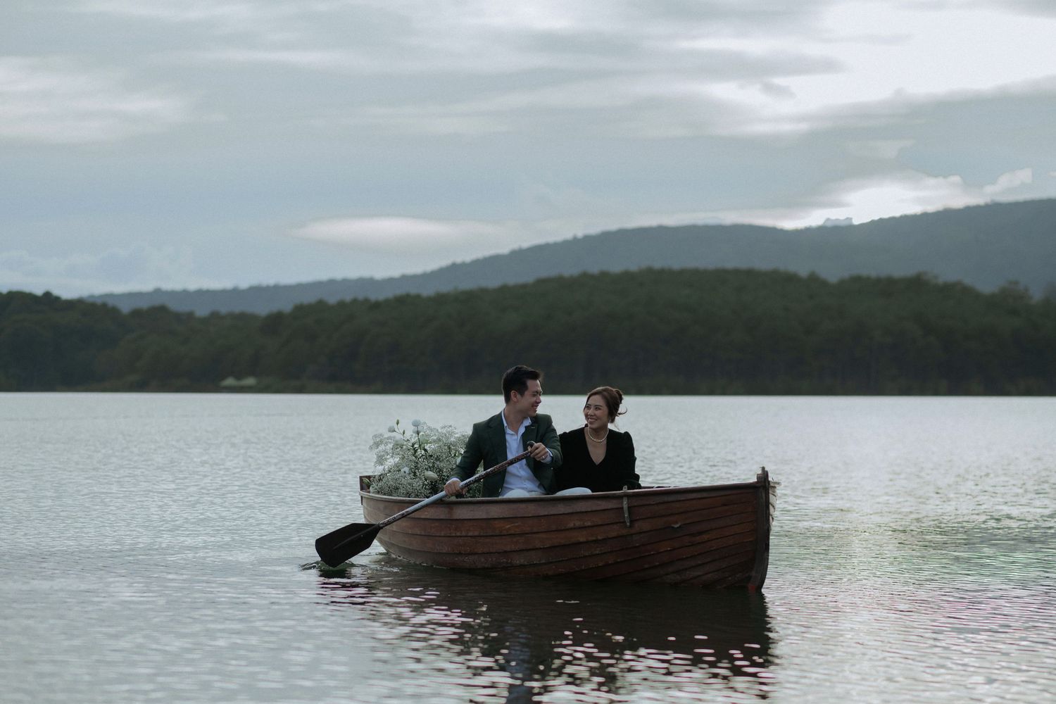 Two people rowing in a wooden boat on a serene lake surrounded by mountains on an overcast day.