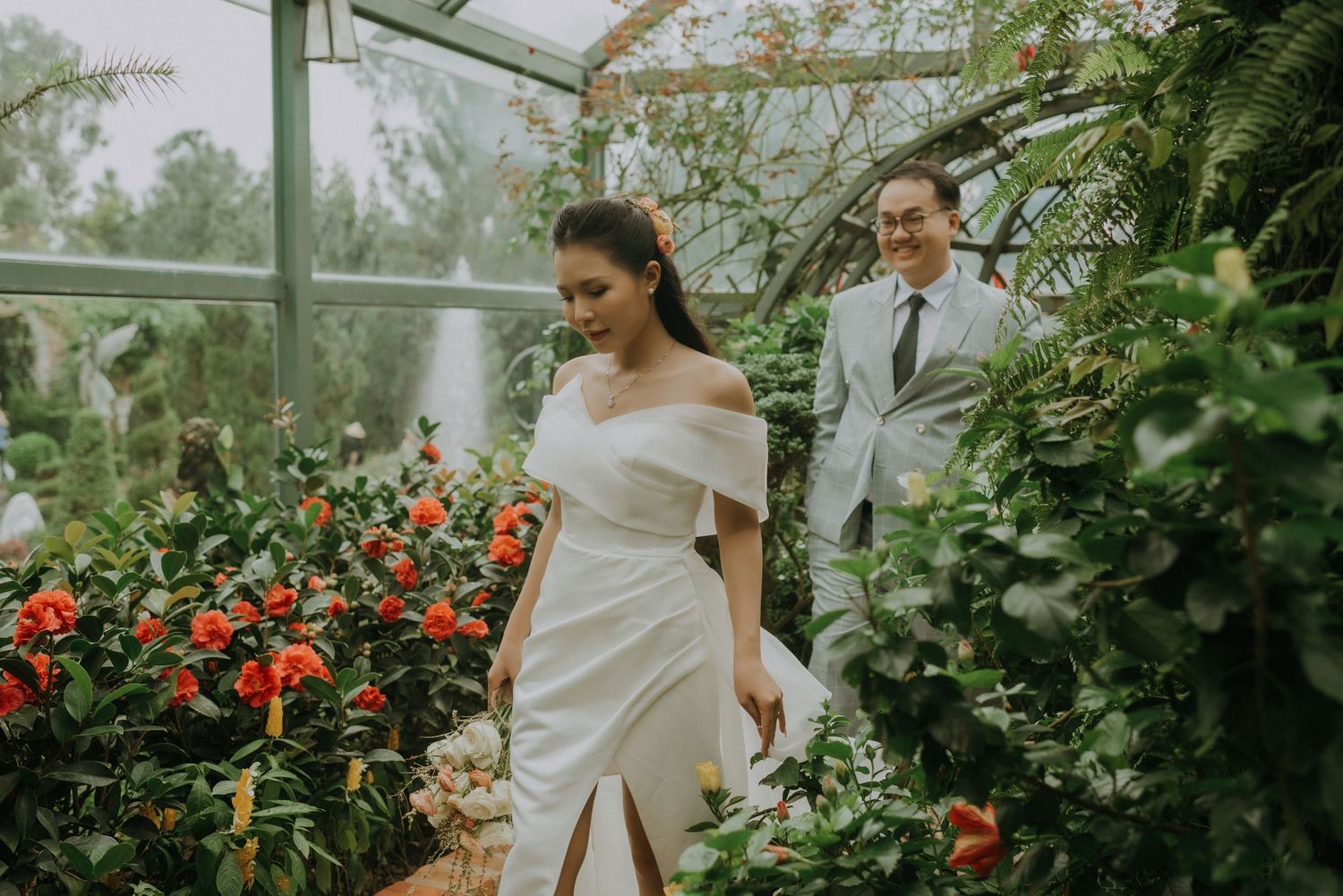 A couple in wedding attire walks through a greenhouse filled with red flowers and lush greenery.