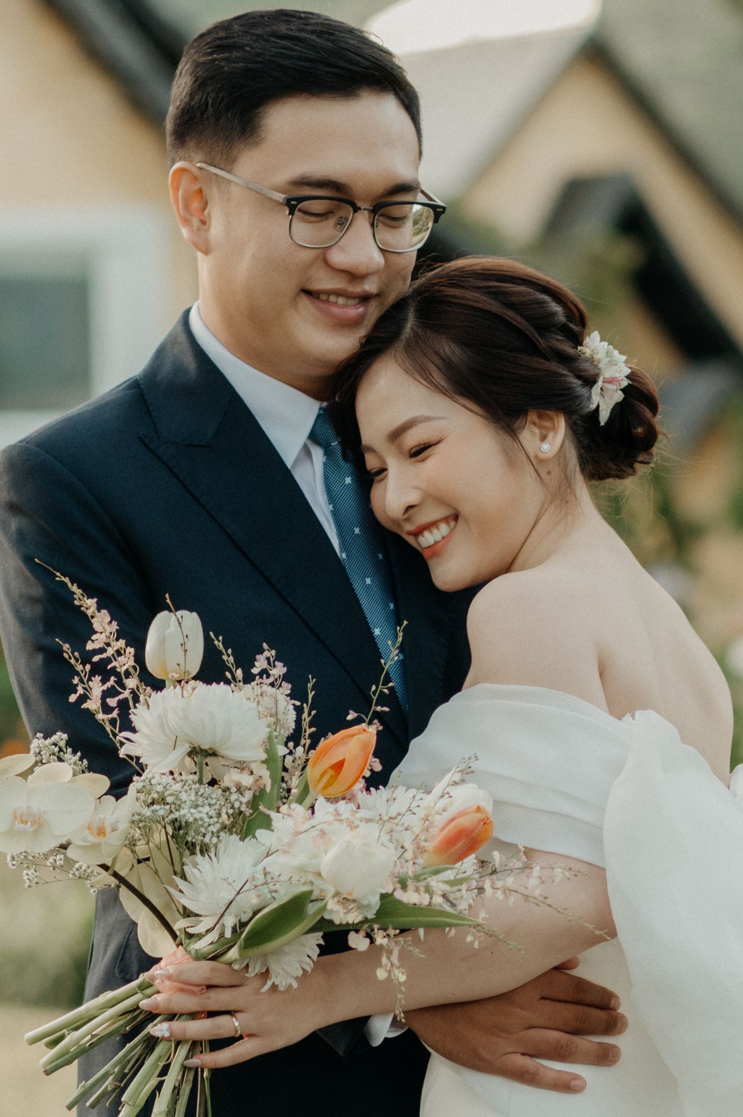 A happy newlywed couple embraces outdoors while holding a beautiful bouquet with white and orange flowers.