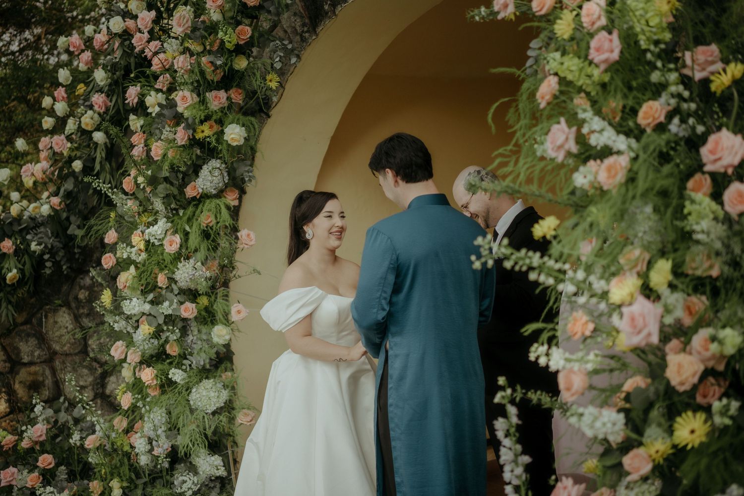 Two people stand under a floral archway covered in pink roses and climbing vines during an intimate outdoor ceremony.