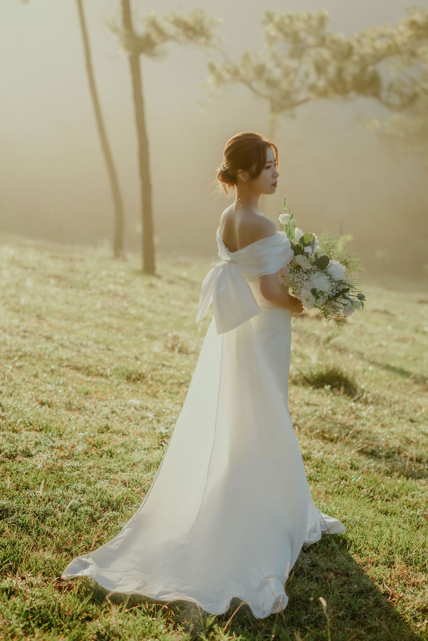 A bride in an off-shoulder white wedding gown holds a bouquet while standing on a misty hillside at golden hour.