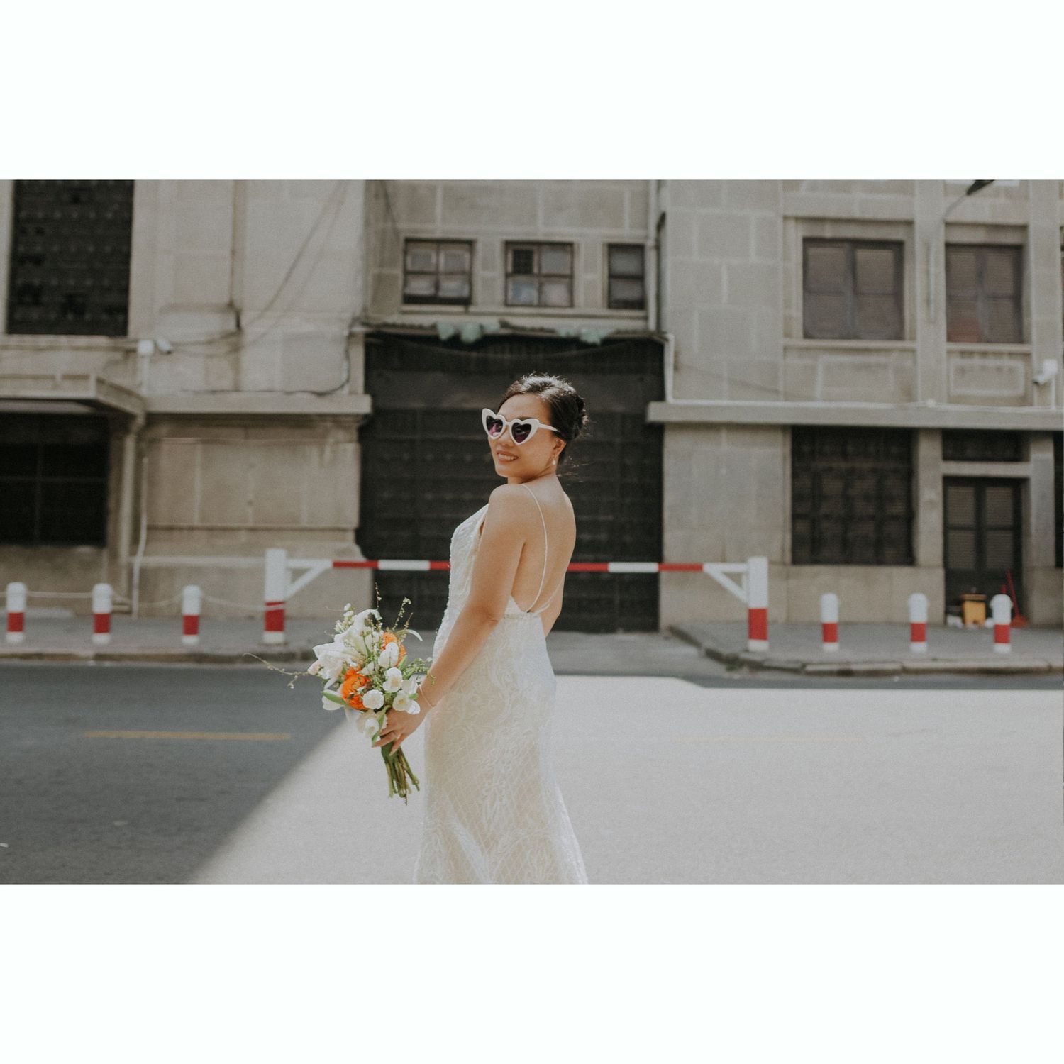 A bride in a classic white dress and sunglasses holds a floral bouquet while standing on a city street.
