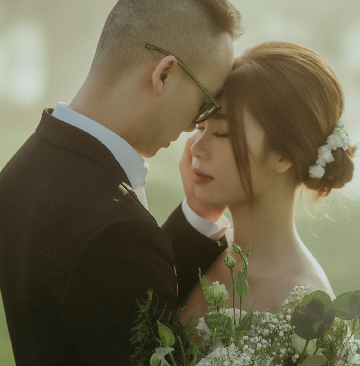 A romantic wedding portrait with a couple in traditional attire holding greenery and flowers, captured in soft lighting.