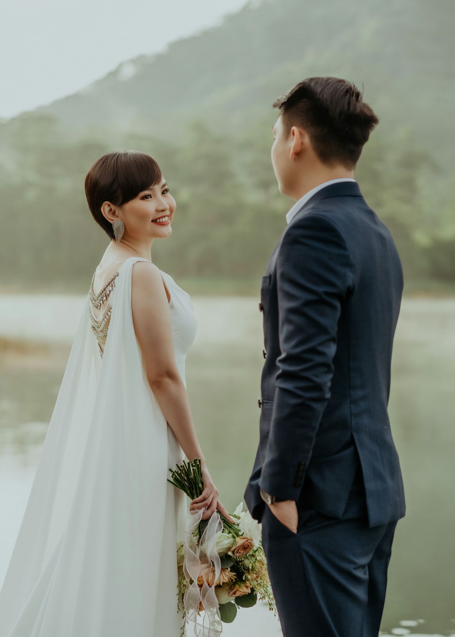 Couple in wedding attire poses together near misty lake with mountains in background at outdoor photo session.