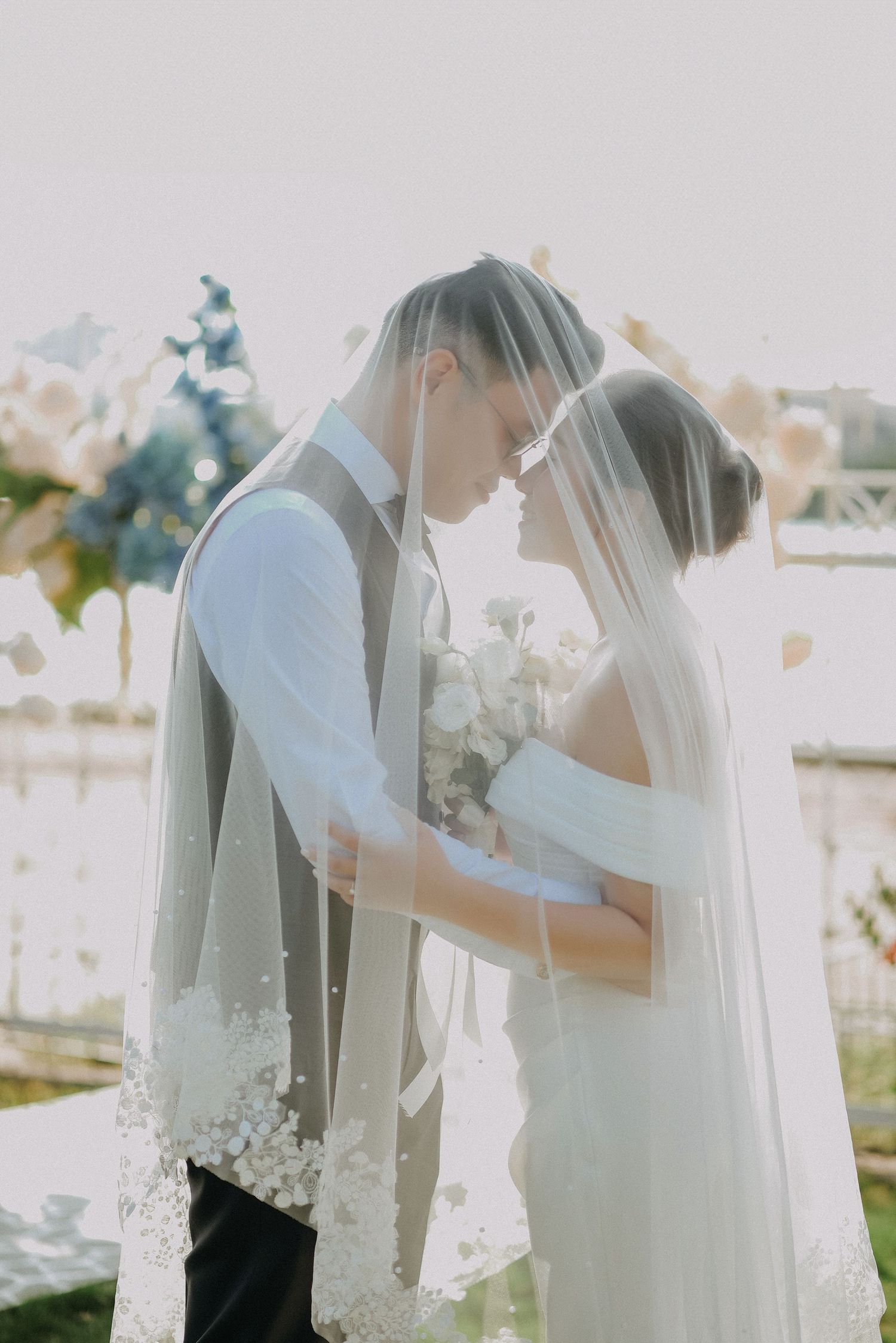 Couple shares an intimate moment under their veils while holding a white bouquet during their wedding ceremony.