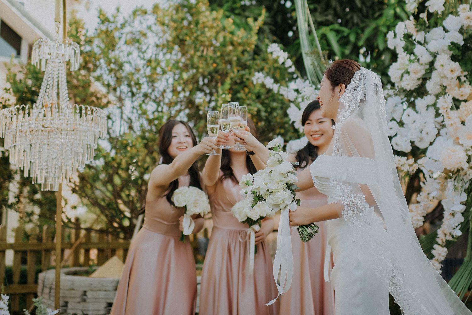 Bridesmaids in pink dresses celebrate with bride among white floral decorations.