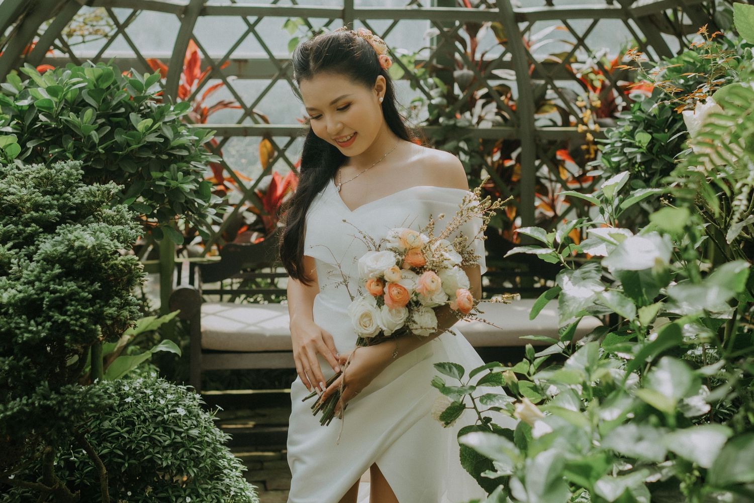 A graceful bride in white dress holds peach-colored bouquet while standing in a lush greenhouse setting.