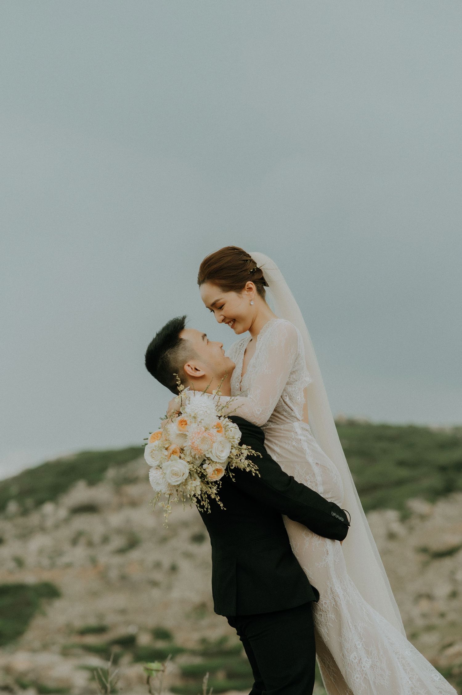 A romantic wedding couple embraces on a hilltop against an overcast sky as the groom lifts his bride holding peach flowers.