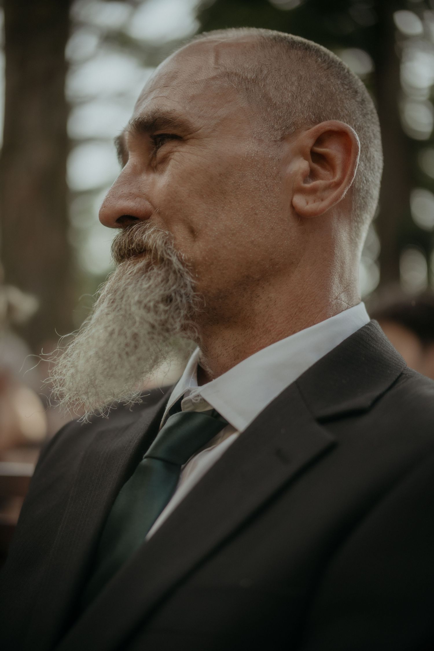 Profile view of older gentleman in dark suit and green tie with long grey beard at formal event.