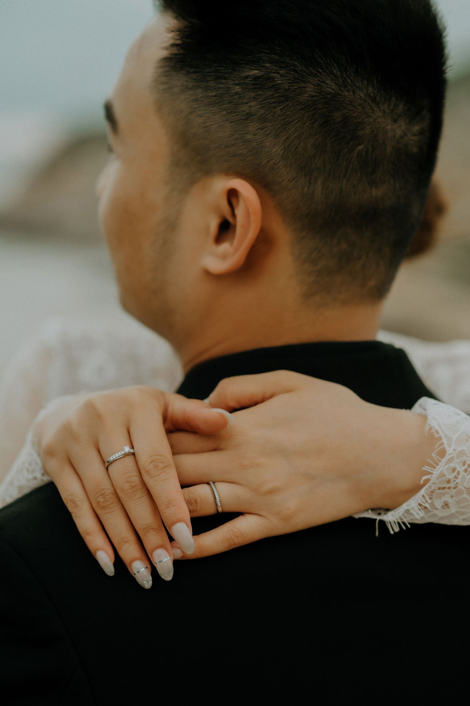A close-up of hands wearing wedding rings embracing, with white lace sleeve detail against a black garment.