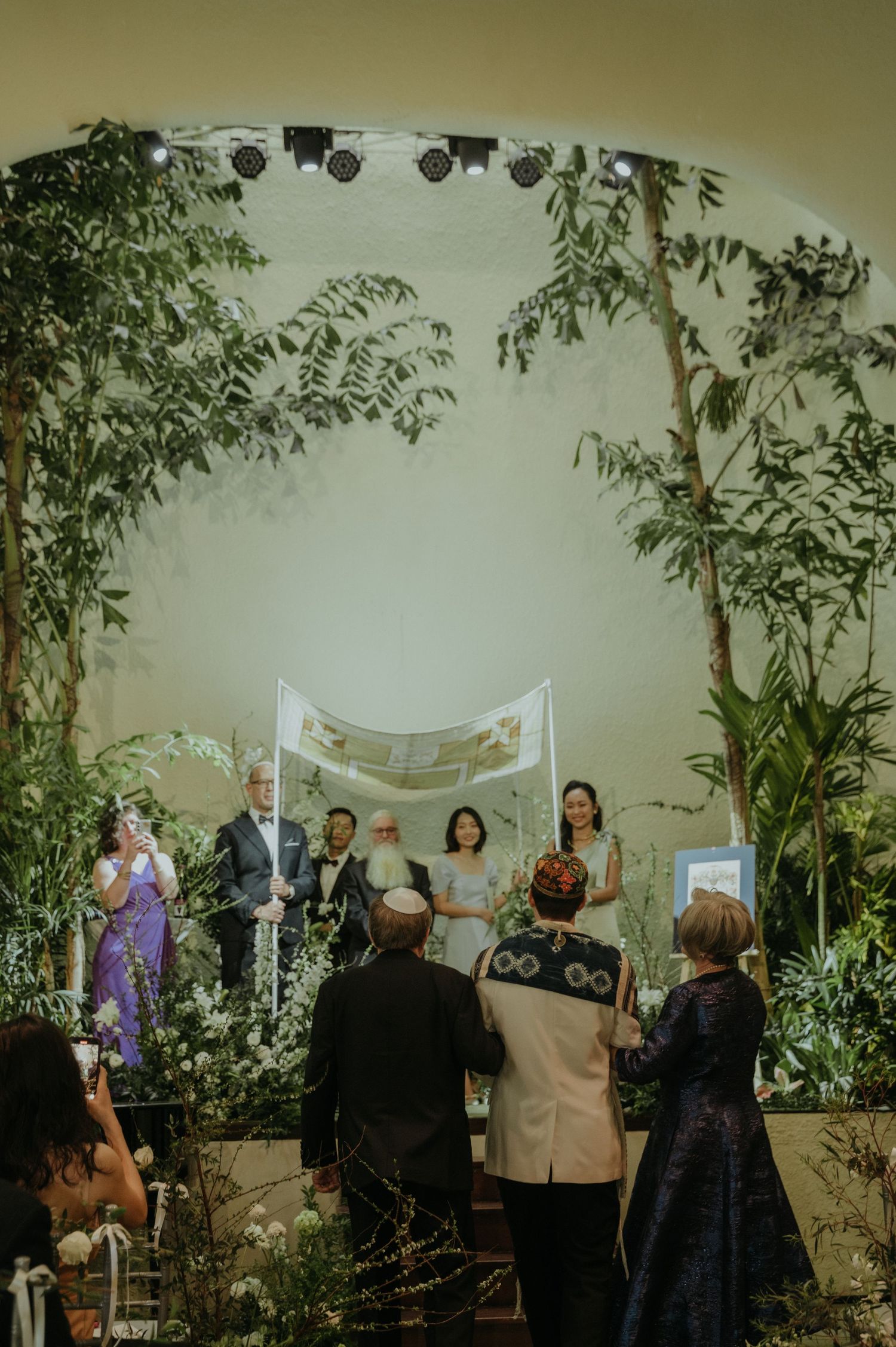 Wedding ceremony space decorated with greenery and white fabric canopy with guests gathered around.