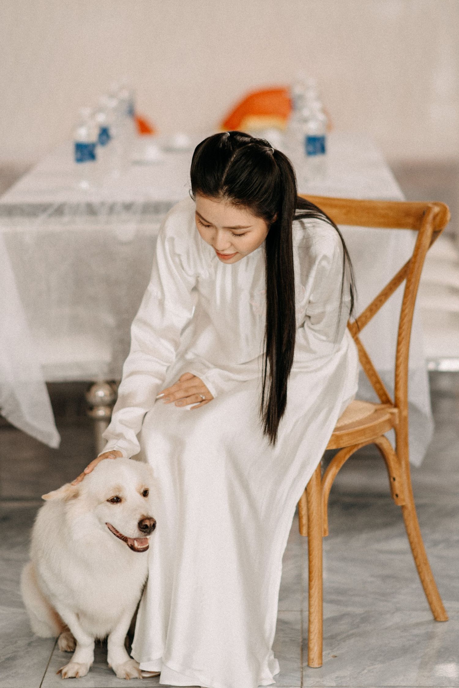 White-robed figure sits in wooden chair petting a fluffy white dog in minimalist interior setting.
