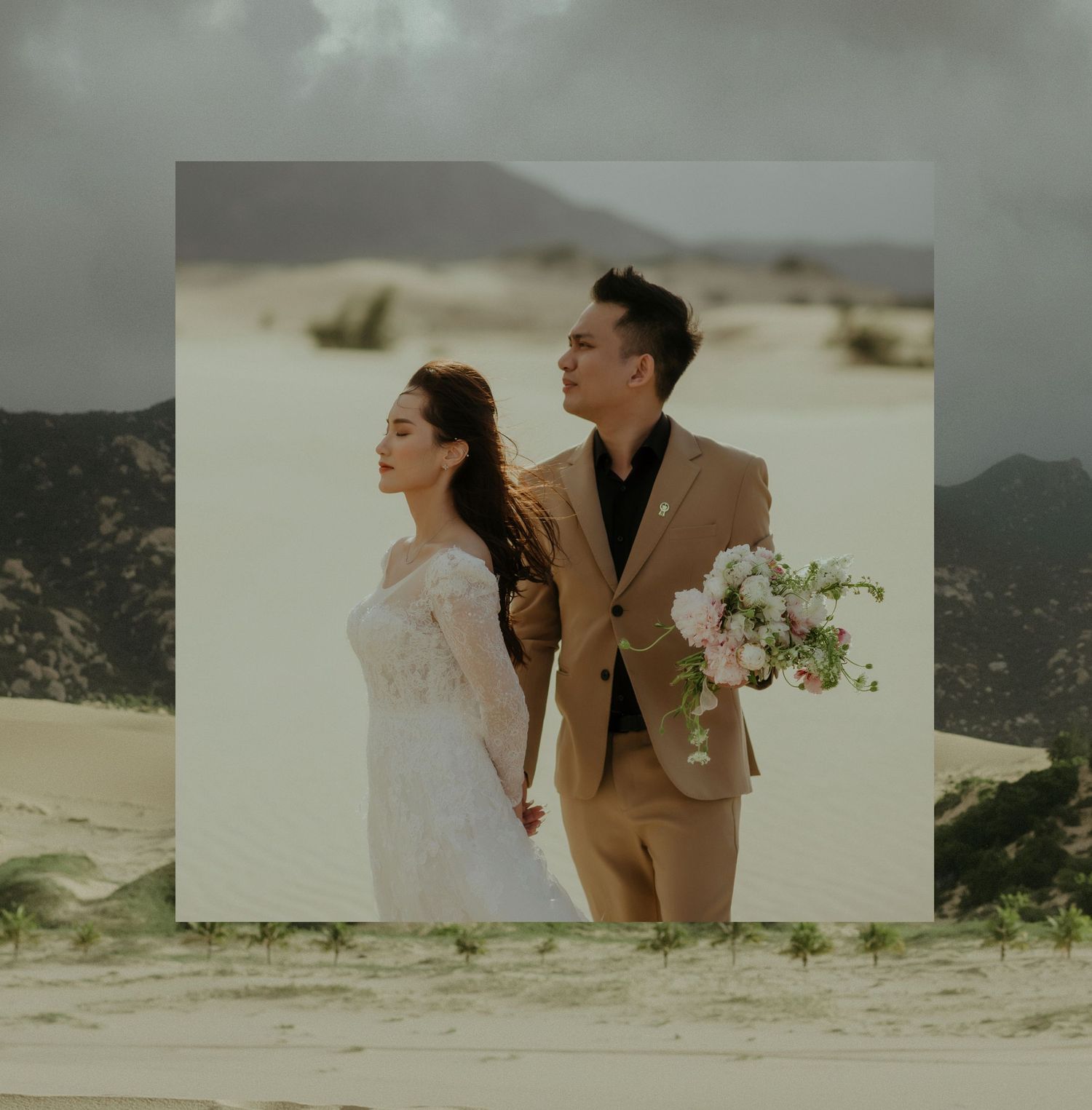 A couple in wedding attire standing together on sand dunes with mountains in background, holding a pink floral bouquet.