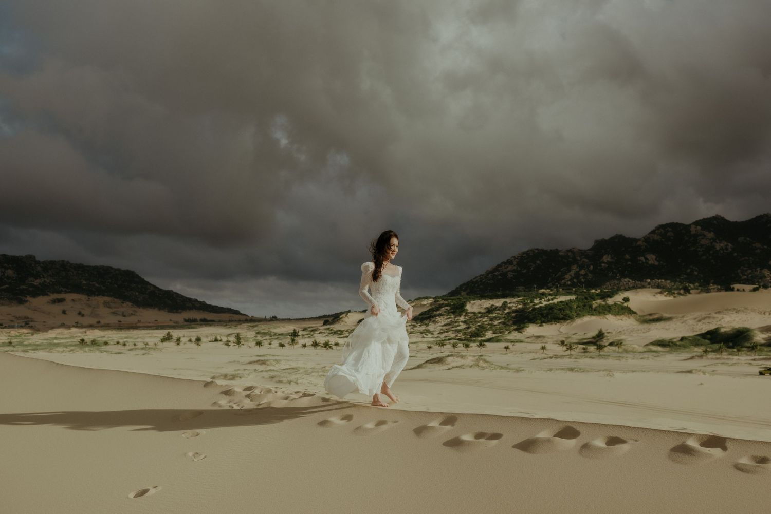 A person in a flowing white dress walks on sandy dunes under dramatic stormy clouds near mountain ranges.