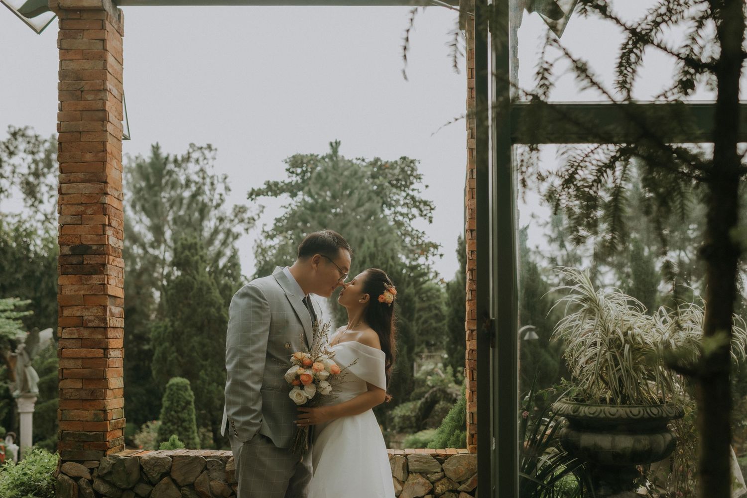 A couple shares an intimate moment in a lush garden setting with brick columns and greenery surrounding them.