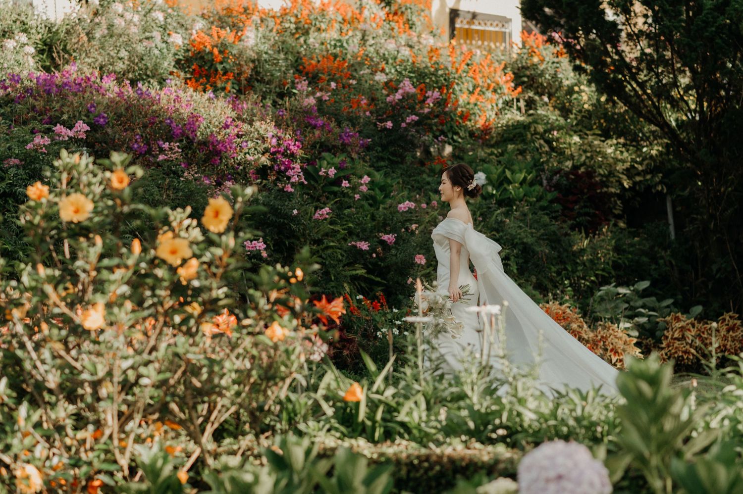 A flowing white gown stands amid a vibrant garden filled with orange, yellow and pink blooming flowers in soft sunlight.