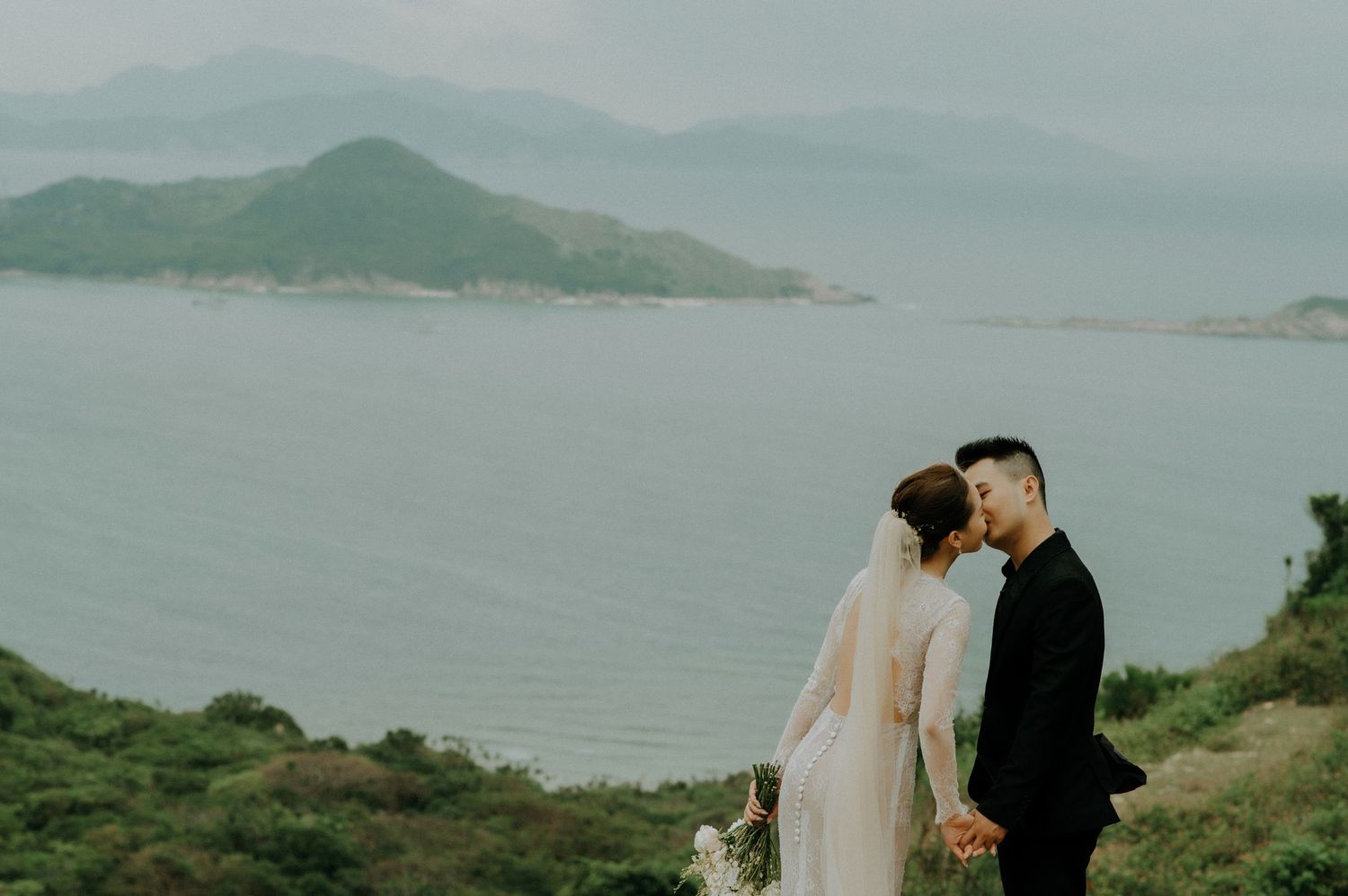 A wedding couple embraces on a scenic coastal mountainside overlooking calm blue waters and misty islands.