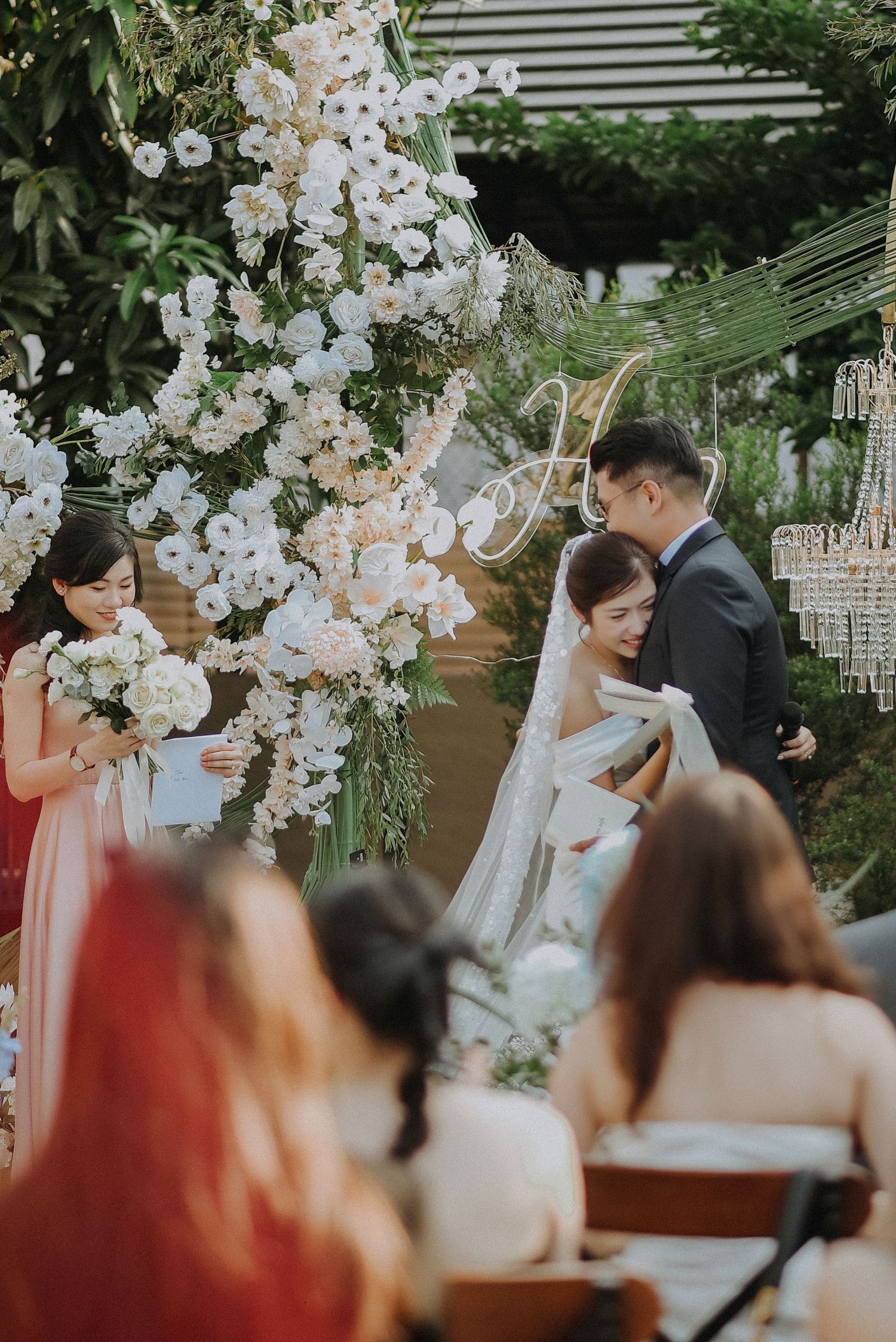 Elegant outdoor wedding ceremony with luxurious white floral arch and crystal chandelier decorations.