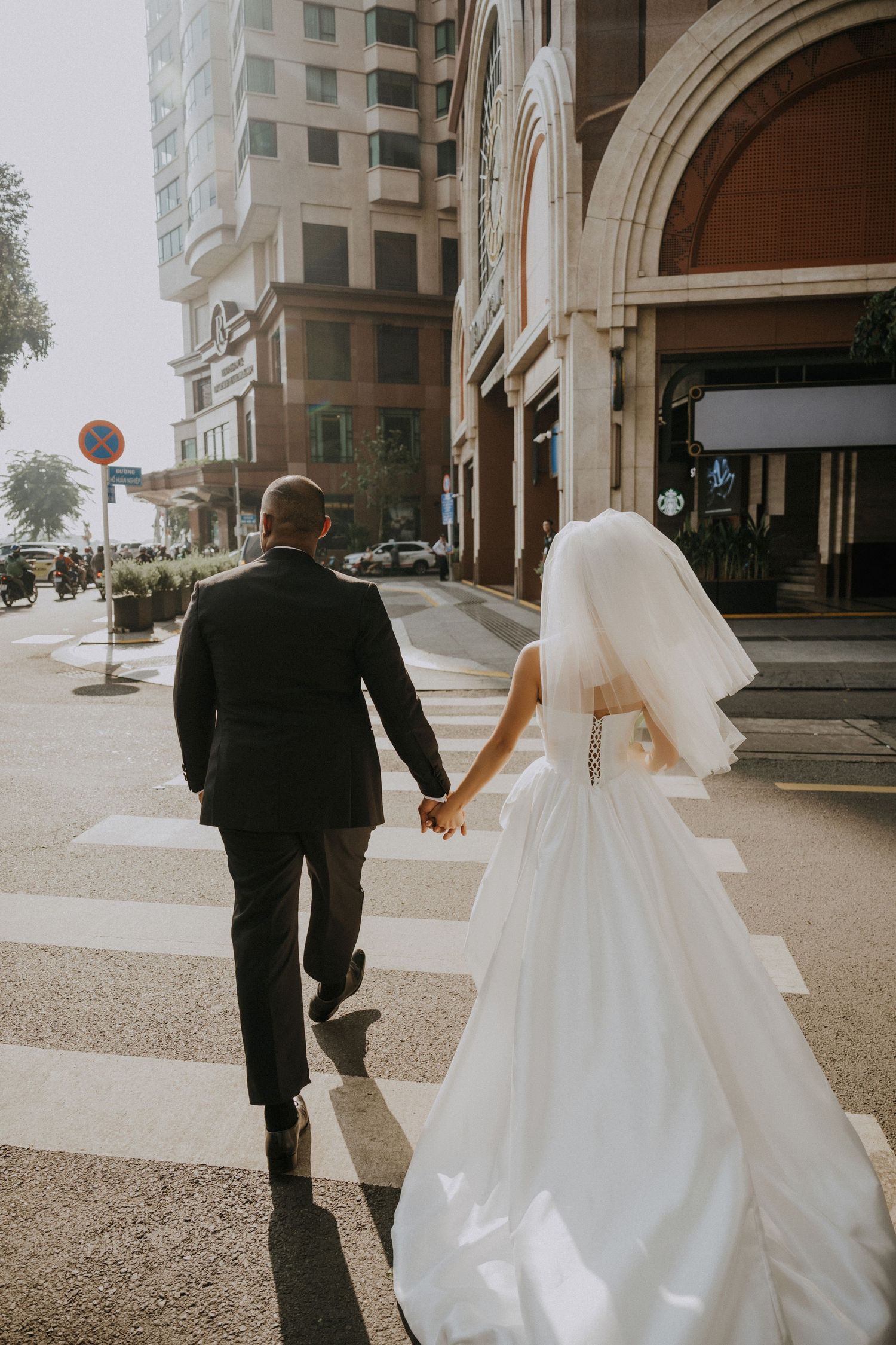 A couple holds hands while walking across a city crosswalk in wedding attire.