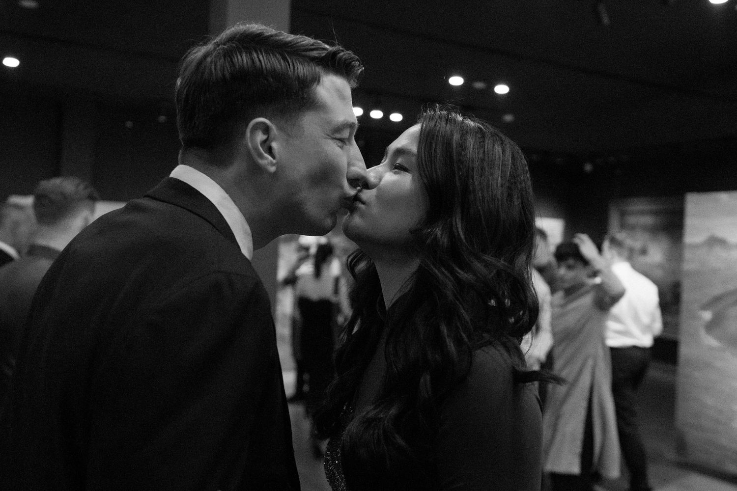 A romantic black and white candid photograph of a couple sharing an intimate moment at a social gathering.