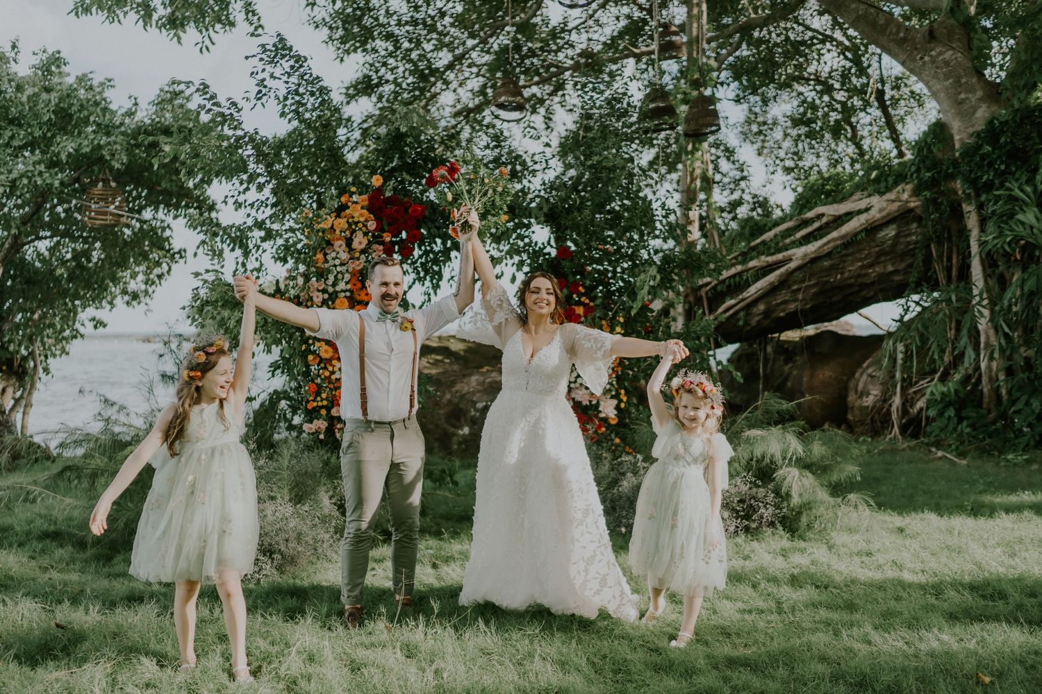 Wedding party jumping joyfully under floral arch in garden setting.