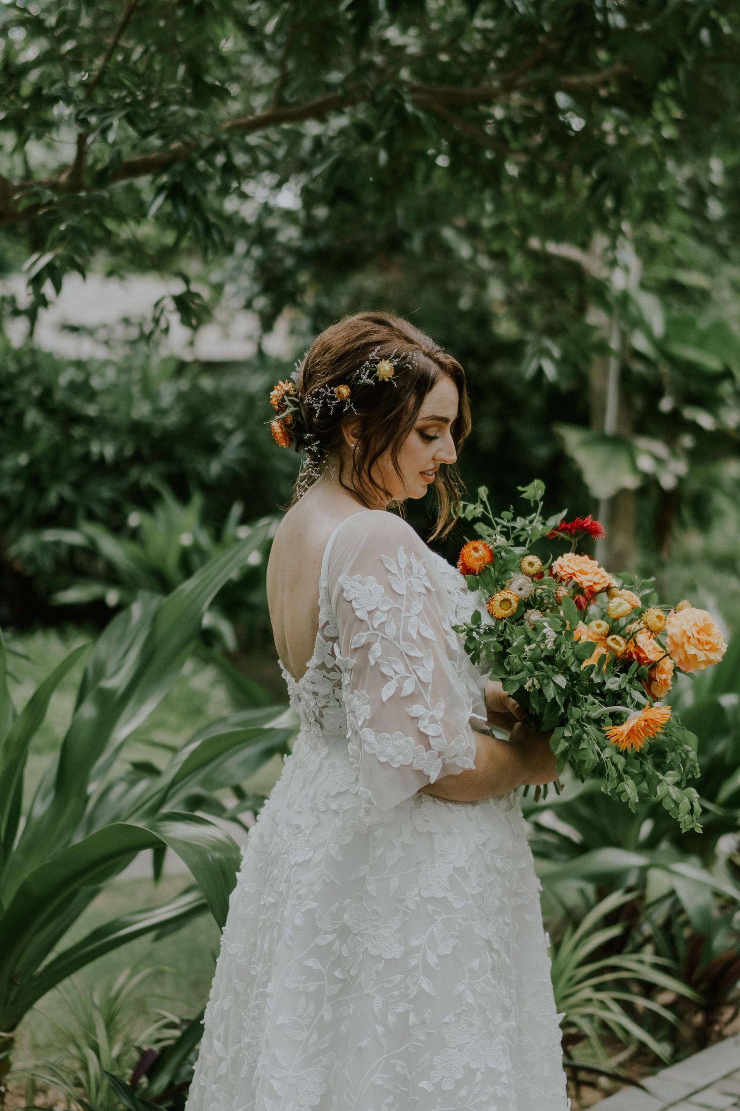 A lace wedding dress and colorful orange floral bouquet stand out against a backdrop of tropical greenery.